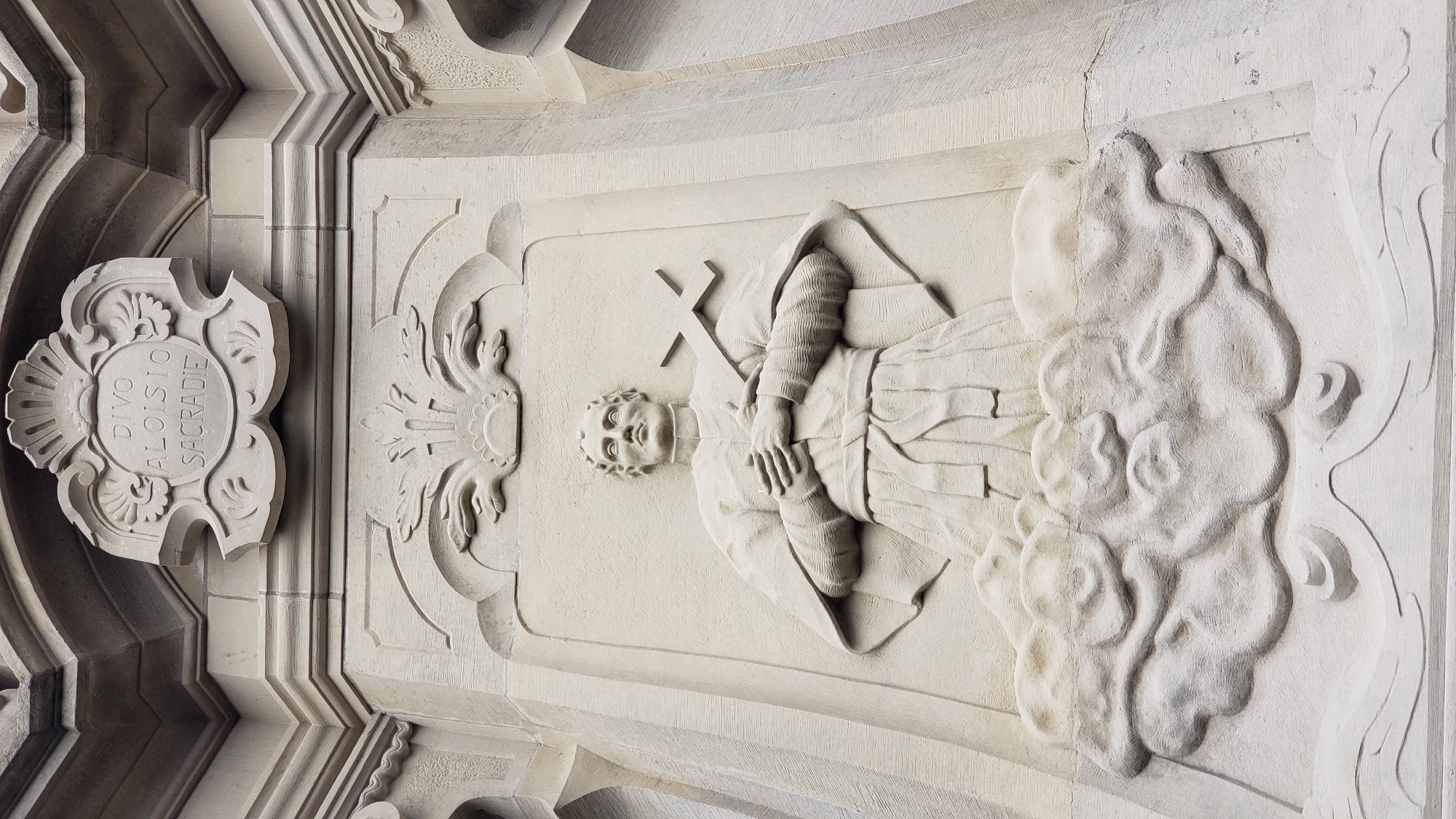 This is a close-up, low-angle exterior shot of a light-colored stone relief sculpture on the side of a building. The sculpture depicts a kneeling male figure dressed in clerical robes, holding a cross with his hands clasped over his chest. The figure is positioned above a sculpted cloud formation, which rests on a decorative base. Above the figure is an ornate crest with Latin text that reads "DIVO ALOISIO SACRAE". The sculpture is located on the side of the St. Nicholas Cathedral in Prešov, Slovakia, a prominent landmark in the city. The stone appears weathered, with some minor discoloration and pitting visible on the surface. The lighting suggests it is daytime, with diffused light casting subtle shadows that highlight the carved details of the relief. The architectural elements surrounding the sculpture are also made of stone and feature decorative mouldings and textures.