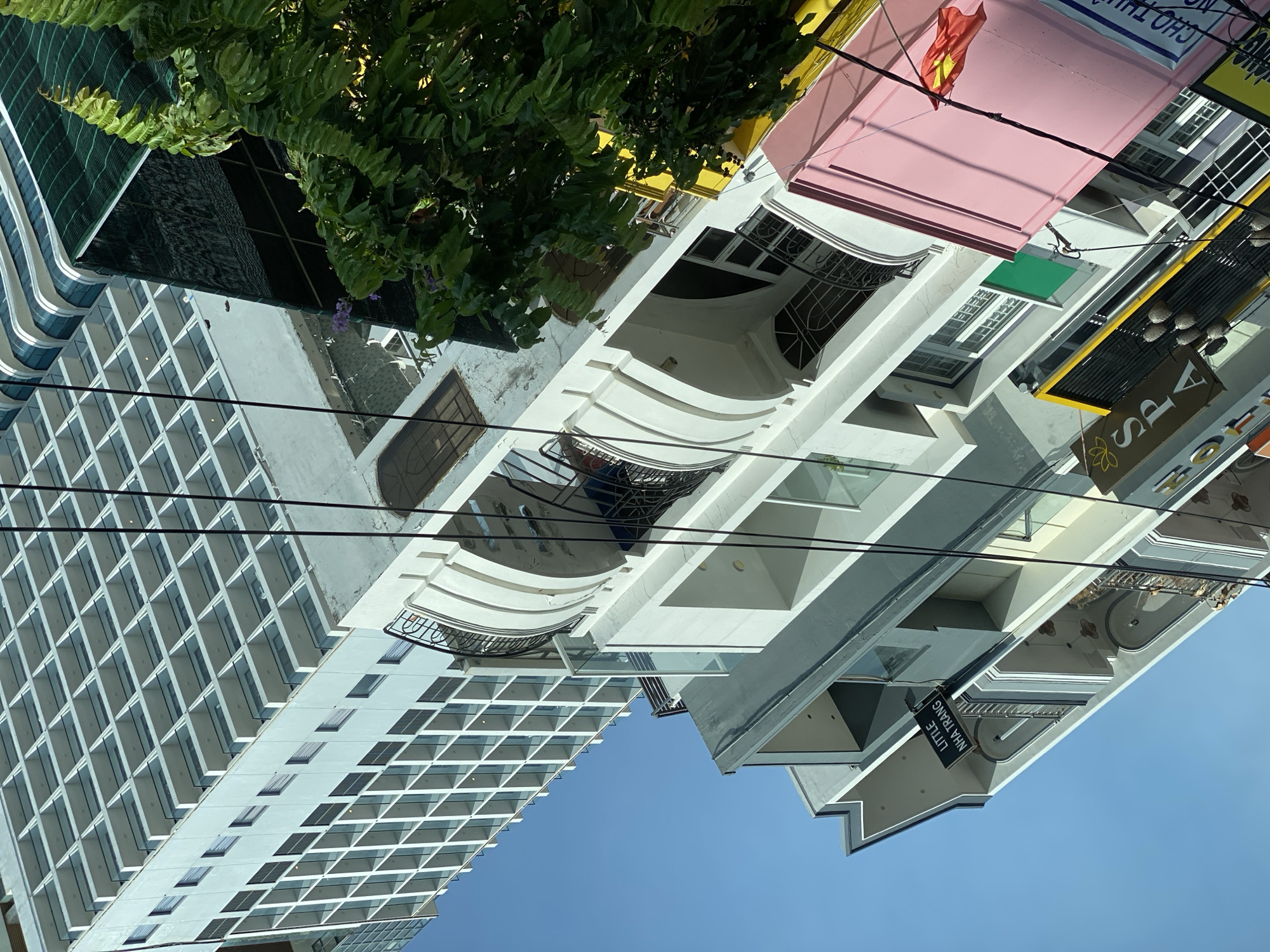 The image displays a street scene in Nha Trang, Vietnam, featuring a multi-story building with a contemporary architectural style. The building is primarily white, with a facade characterized by repetitive balconies and windows. Above this structure, a portion of a much taller building is visible, its upper floors showcasing a grid-like pattern of balconies or windows. Electrical wires crisscross the frame, indicating urban infrastructure.
Several smaller buildings are situated in the foreground and to the right, exhibiting a mix of architectural elements. One building features a prominent pink facade on its upper story, with a Vietnamese flag attached to its exterior. Below this, a bright yellow sign with the word "SPA" in a floral font is visible, along with other signage that appears to be in Vietnamese. Another sign on a gray building reads "LITTLE NHA TRANG." A lush green tree with dense foliage is present on the right side of the image, partially obscuring some of the buildings. The sky is a clear, bright blue. The perspective is angled, looking upwards at the buildings, suggesting the photo was taken from ground level or a lower vantage point. No people are clearly discernible in the image.