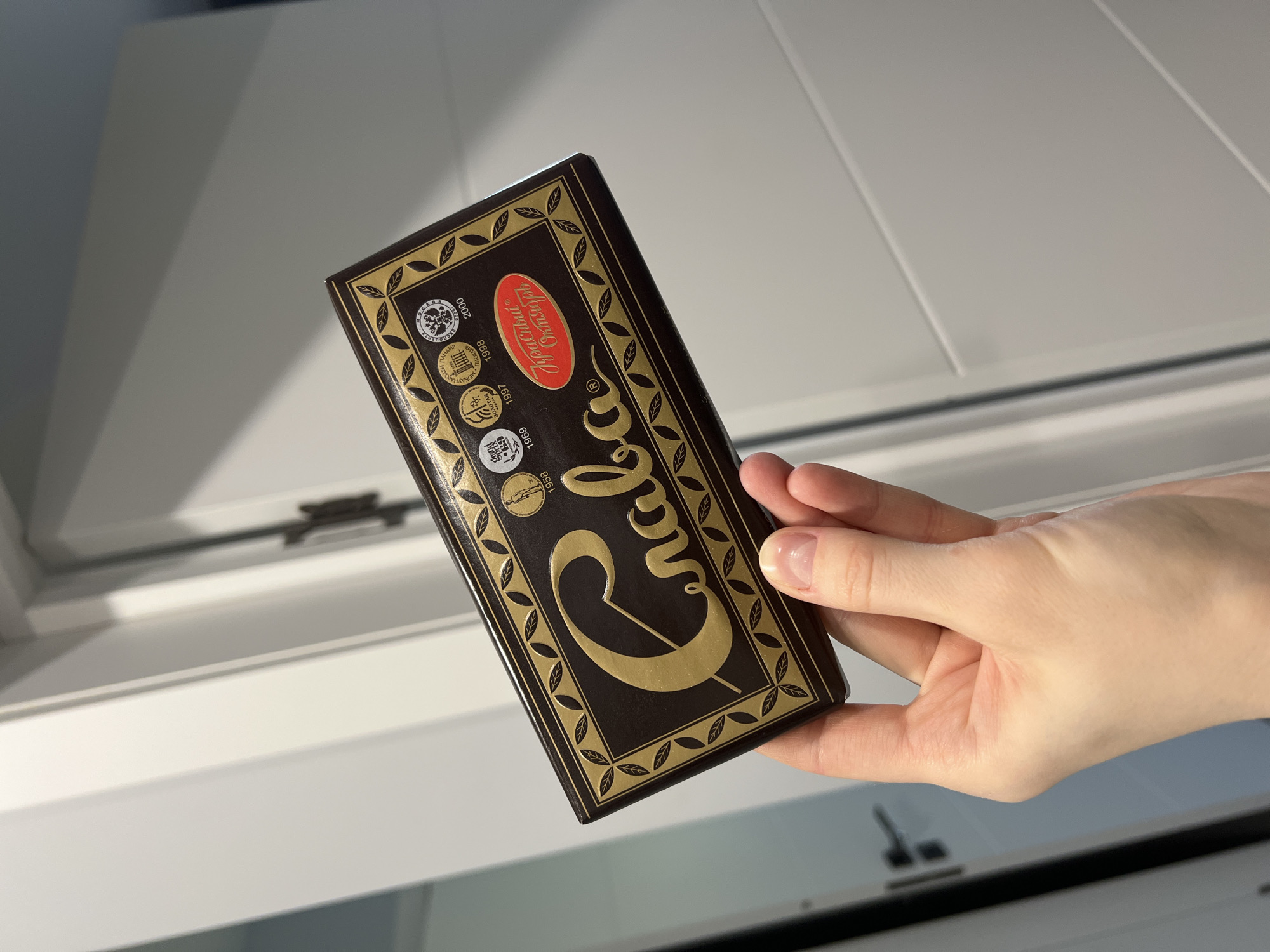 A hand holds a rectangular bar of chocolate with gold lettering on a dark brown background. The chocolate is likely in a room with white walls and a window frame. The packaging features the Cyrillic word "Grilyaж" (Grilyazh) and several round symbols with years inscribed within them.