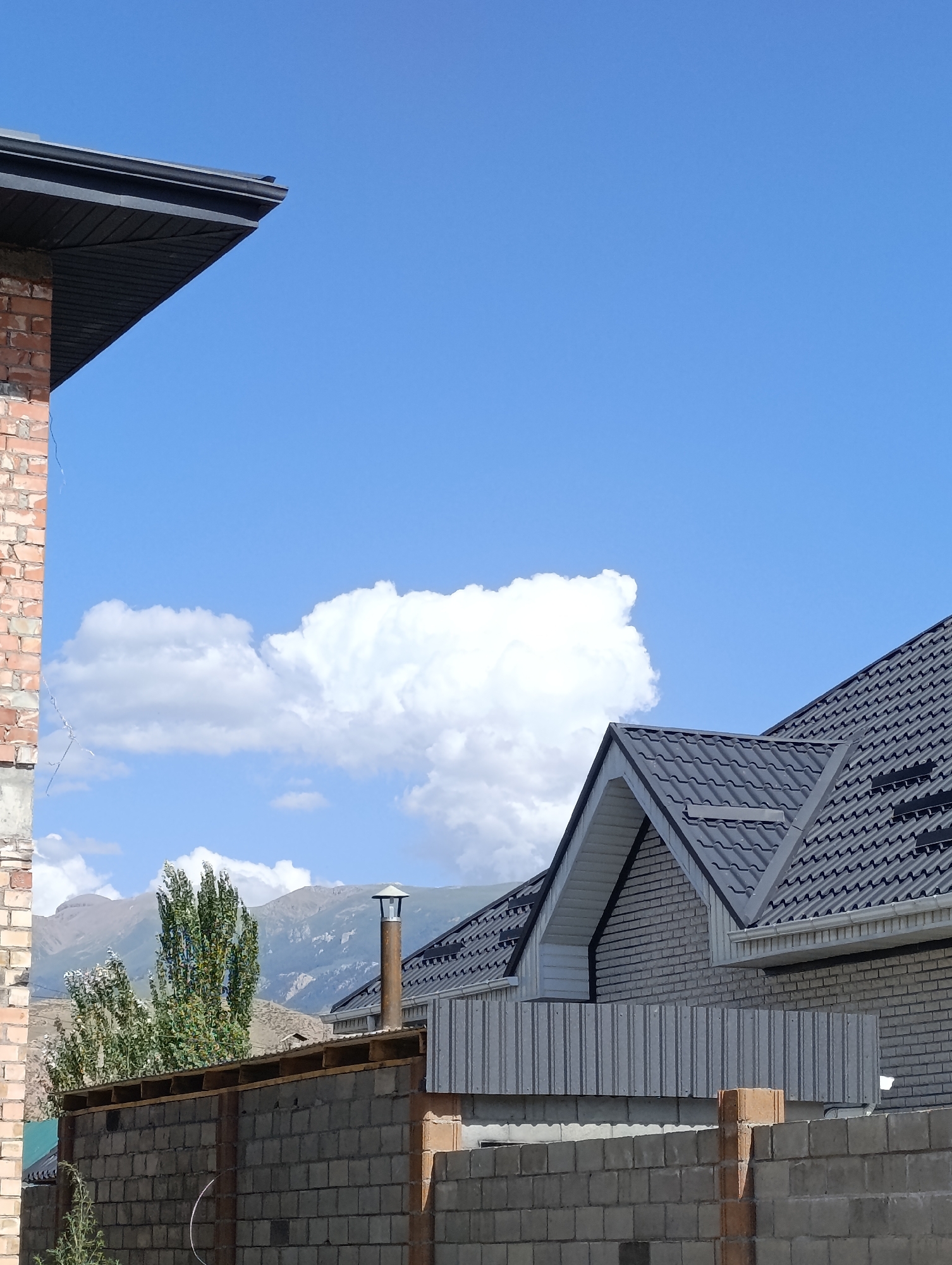 This is a medium-angle, outdoor, daytime photograph taken in Naryn City, Kyrgyzstan. The image depicts a partially constructed residential area with elements of traditional and modern architecture. In the foreground, a fence made of concrete blocks is visible. Beyond the fence, there are houses under construction with gray tiled roofs and white brick exteriors. A tall chimney with a metal cap rises from one of the houses. In the background, lush green trees stand against a backdrop of mountains. The sky is a vibrant blue with scattered white clouds. The lighting suggests a sunny day.