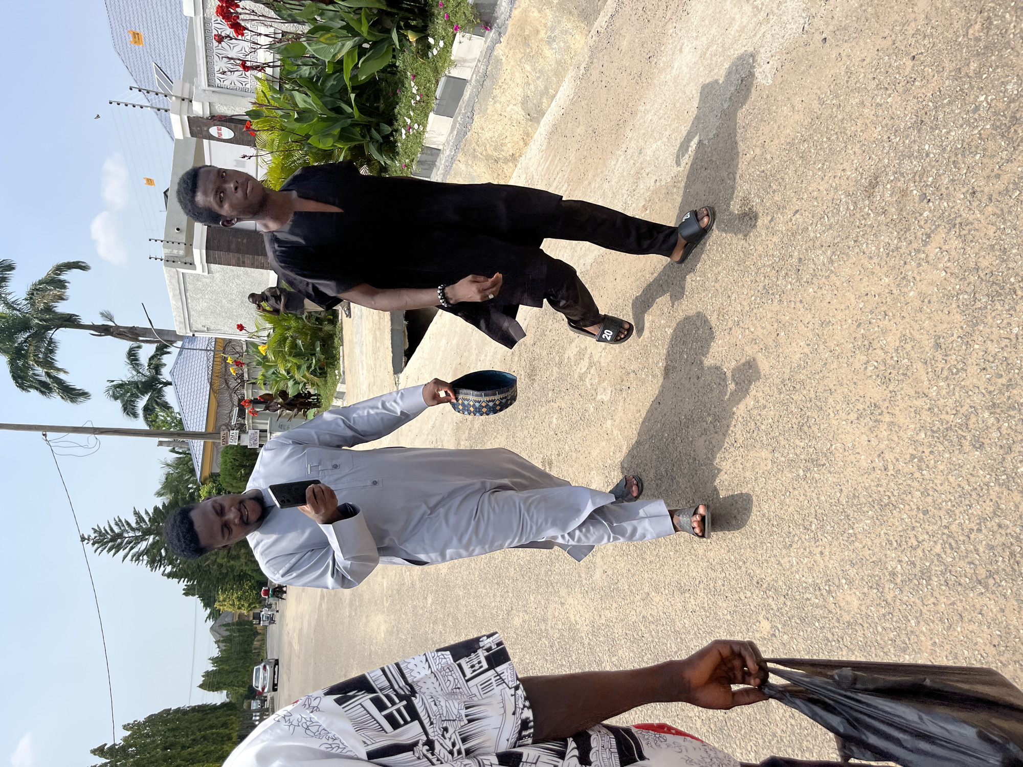 Two men are pictured in an outdoor setting. The man in the foreground, on the left side of the image, is wearing a light grey traditional outfit and sandals. He is holding a black smartphone in his right hand and a patterned cap in his left. He appears to be smiling and looking towards the camera.
The second man, on the right side of the image, is wearing a black traditional outfit and black slides with the number "20" visible on one of them. He is looking towards the camera with a neutral expression. He has a bracelet on his left wrist.
Behind the men, there is a paved pathway, lush green vegetation with red flowers, and a building with a white fence. Palm trees are also visible. The scene suggests it is daytime with clear skies. The location context provided is Abuja, Nigeria. The overall mood appears casual and friendly.