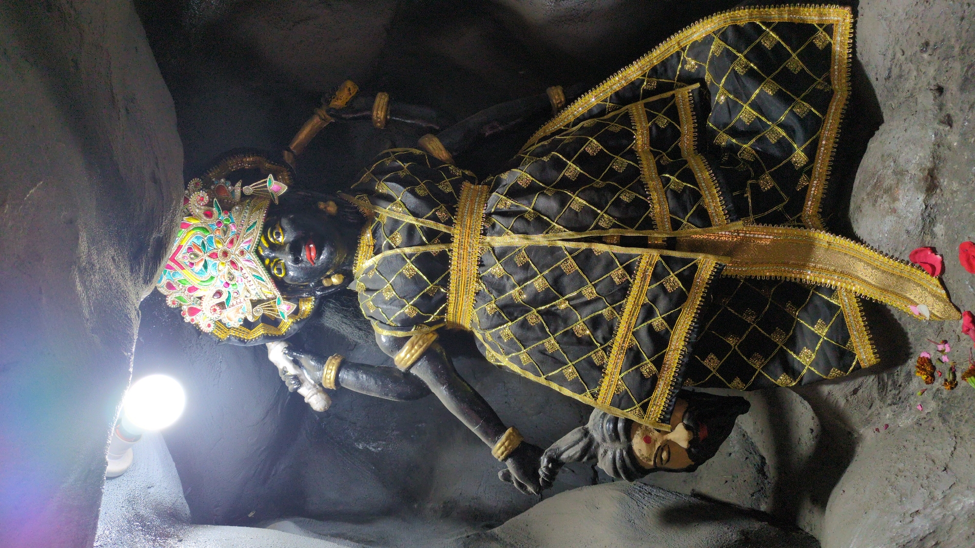A prominent statue of a Hindu goddess, characterized by her dark complexion, vibrant multi-colored headdress, and intricate black and gold patterned attire, dominates the frame. Her piercing yellow eyes and red lips are distinctive features. She is adorned with gold bangles on her wrists. Below her feet lies a prostrate figure, typically identified as Shiva, with a human-like face, mustache, and red mark on his forehead, a common depiction in Kali iconography.
The setting appears to be a cave or a rock-cut shrine, with rough, dark, and uneven rock formations forming the walls and ground. A bright, bare light bulb on the lower left casts strong illumination on the scene. Scattered red and pink flower petals are visible on the ground around the deities, indicating offerings or religious rituals. The image captures a devotional scene, common in temples or sacred sites. No text is visible in the media file. The location context suggests this is a shrine in Bhagsu, India.