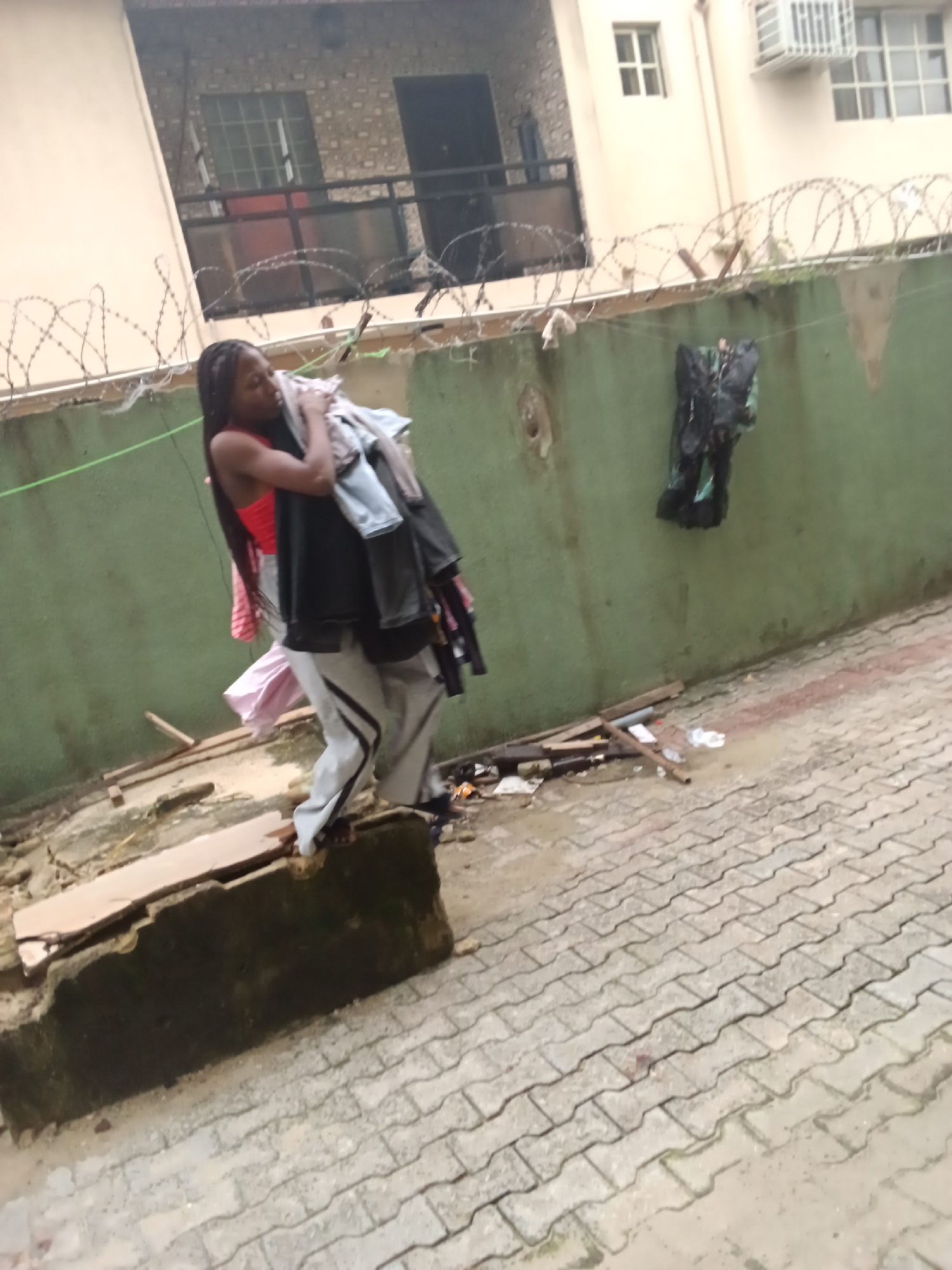 A medium-angle full shot captures a young individual, likely female, with long dark braided hair, in an outdoor residential or semi-residential area of Ado, Nigeria. The person, wearing a red sleeveless top and light grey sweatpants with a black stripe, is carrying a substantial pile of various clothes, including a black jacket or shirt, a light blue item, and a pink garment, clutched against their chest with both arms. They appear to be stepping or standing on a low, broken concrete structure or ledge, with wooden planks and other debris scattered around the uneven, dirty ground immediately in front of them.
The ground beyond the debris consists of interlocking concrete pavers, which appear damp and somewhat dirty. In the mid-ground, a green-painted concrete wall runs horizontally across the frame, with sections of its paint chipped away and signs of dirt or mold. A thin green string or wire is stretched along the wall, from which a dark, possibly black, piece of clothing or fabric is hanging to dry or simply placed. Above this wall, multiple strands of barbed wire are installed, stretching across the scene.
In the background, a light-colored, possibly cream or beige, building with multiple windows and a balcony is visible. One window features a metal gridded air conditioning unit. The overall impression is one of a lived-in, somewhat cluttered urban environment.