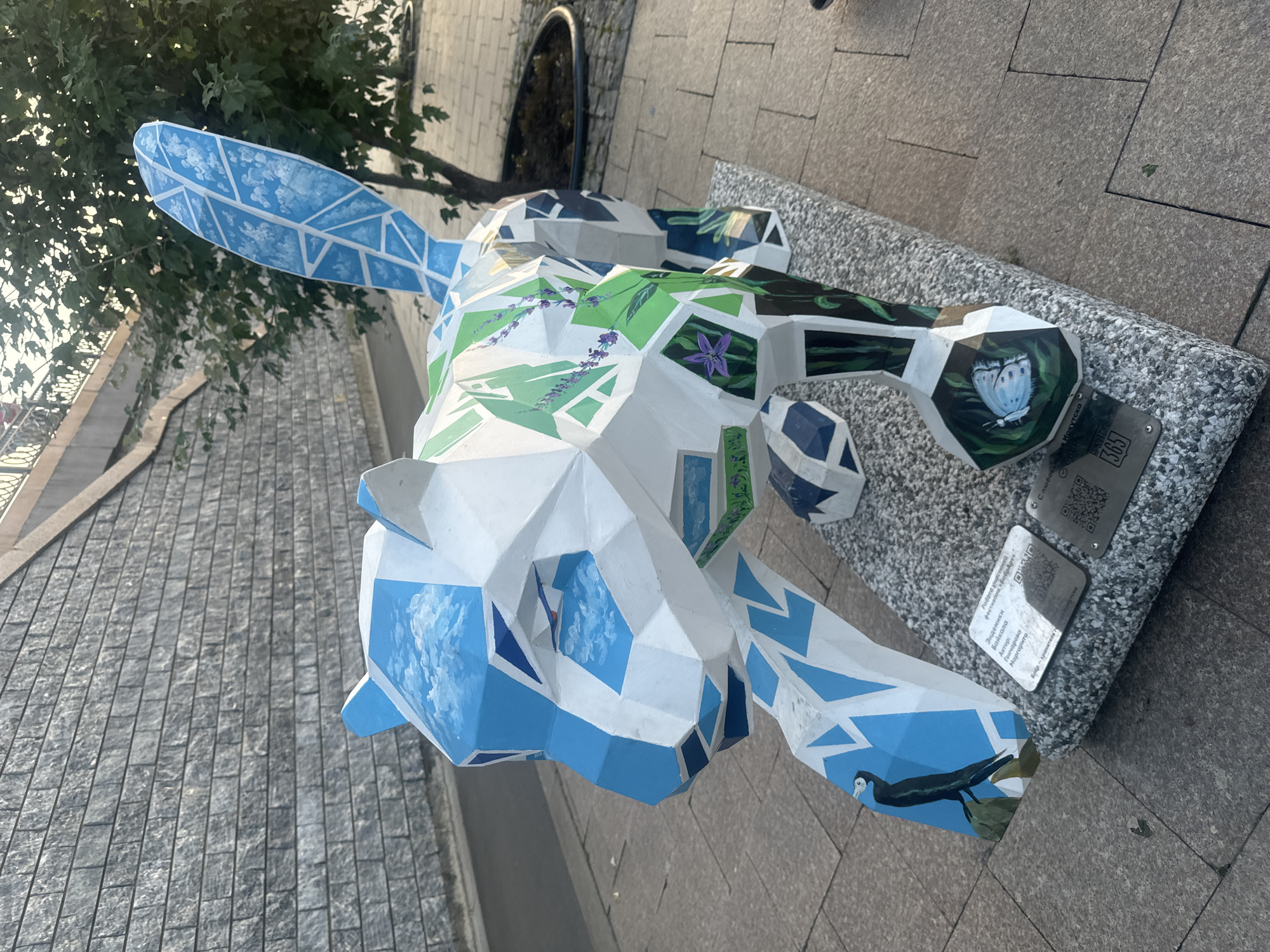 This is a photograph of an art installation in Irkutsk, Russia. The main subject is a polygonal sculpture of a stylized animal, possibly a fox or a lynx, painted with various natural scenes. The sculpture is predominantly white with sections painted blue, green, and with depictions of clouds, lavender, a bird, and a butterfly. It stands on a grey, speckled base on a paved walkway.
To the left of the sculpture is a stone retaining wall made of evenly cut grey blocks. A paved walkway extends in front of the wall and around the sculpture. In the background, a tree with green foliage is visible, and beyond that, a section of a railing and a slope leading upwards, suggesting a riverside embankment or a bridge.
On the base of the sculpture, there are two metallic plaques with text and QR codes. One plaque contains Cyrillic text and appears to be an artist's statement or information about the artwork. The other plaque has a QR code and a number "365".
The lighting suggests it is either early morning or late afternoon, with soft, warm light casting shadows. The environment appears to be an outdoor public space. No people are clearly visible in the image, though the presence of a railing and walkway indicates a public area.