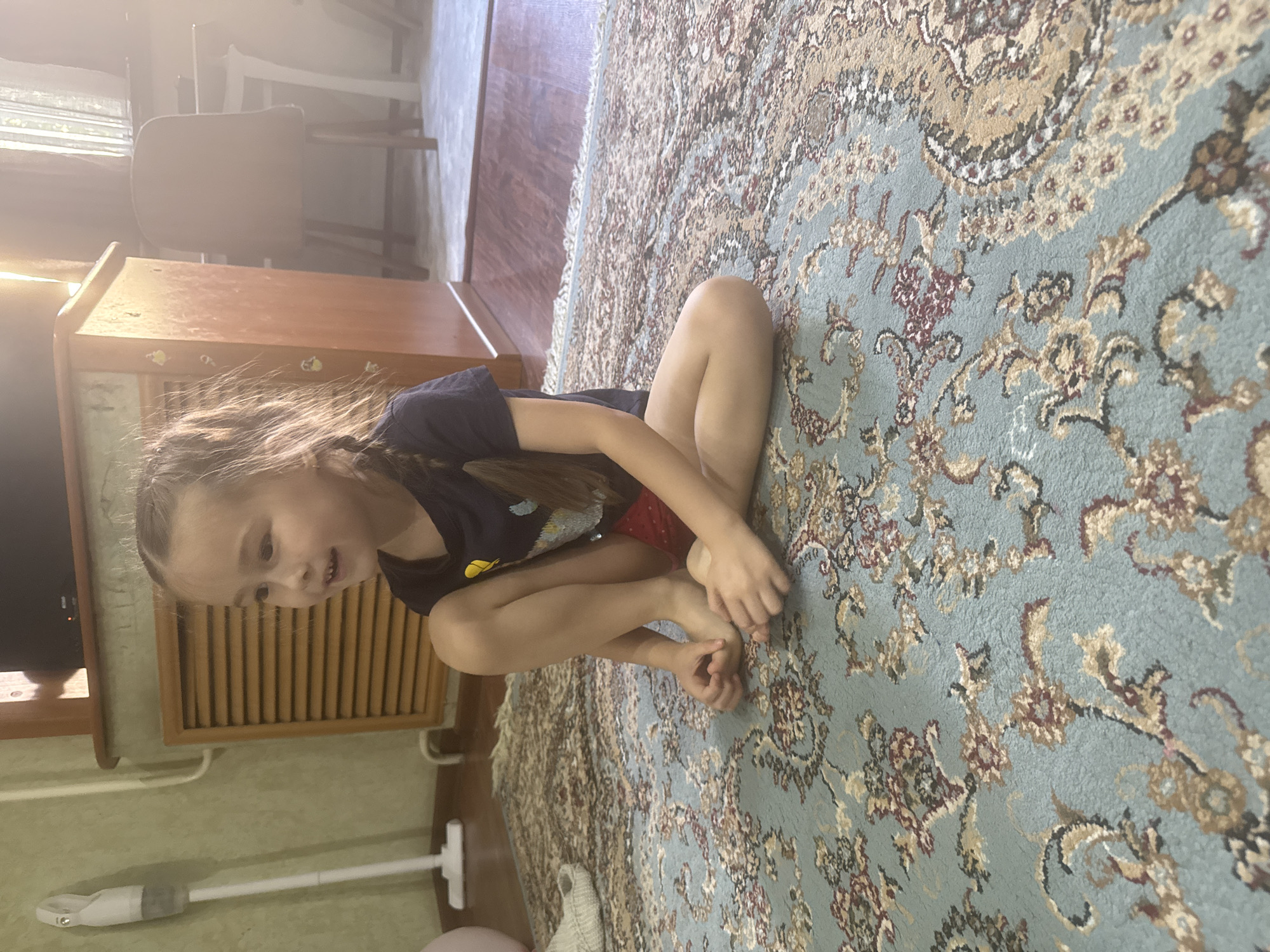 A young girl sits cross-legged on an ornate, light blue rug with floral patterns in a room. She wears a dark short-sleeved shirt and red shorts. The room includes wooden furniture, a window, and a partially visible vacuum cleaner. The location is Tashkent, Uzbekistan.