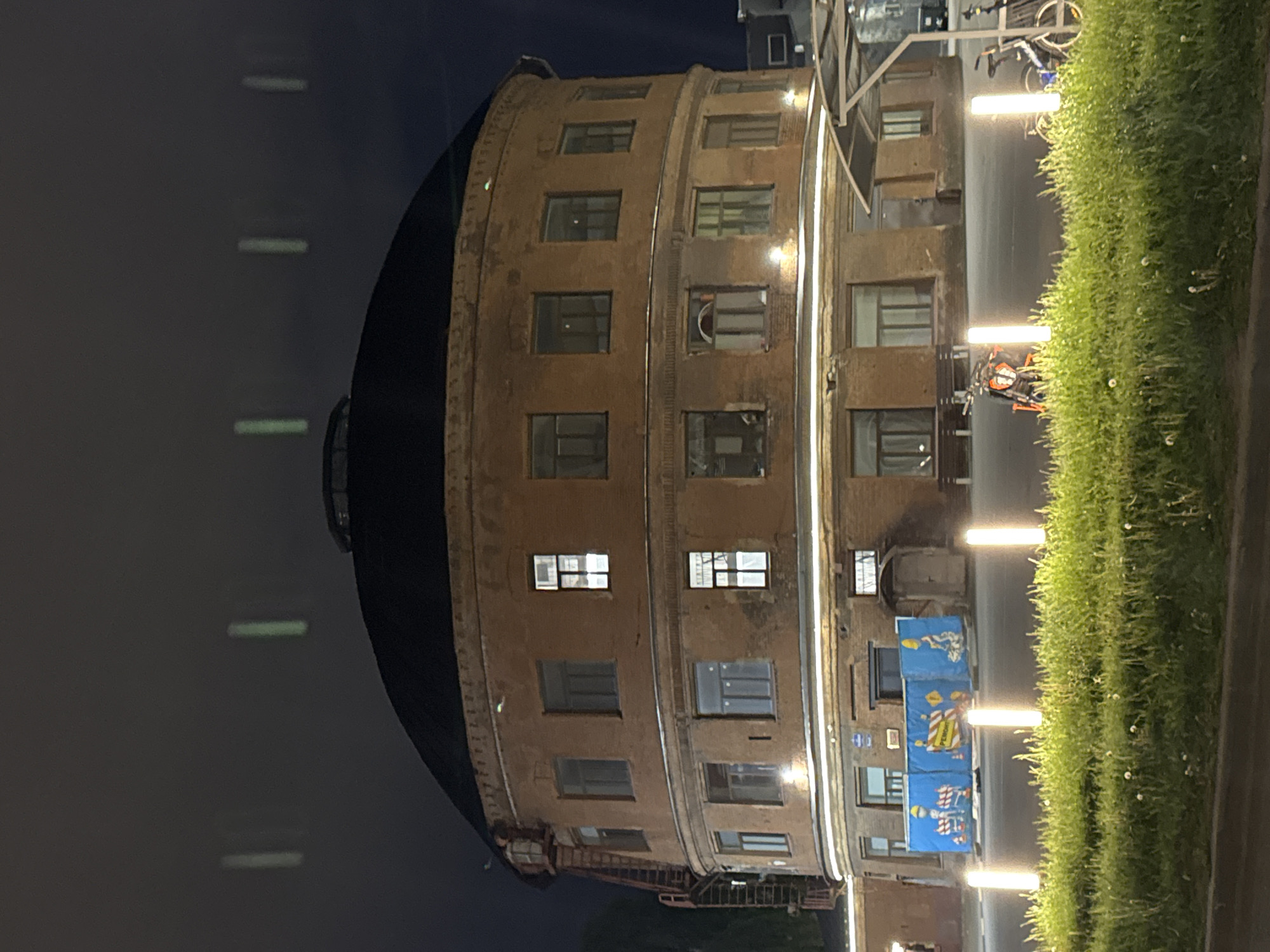Here is a description of the image:
The image shows the Water Tower on Shpalernaya Street in Saint Petersburg, Russia, at night. The tower is a large, cylindrical, brick structure with multiple rows of windows and a dark, domed roof. A few vertical lights are positioned between the building and the edge of the image. The lower half of the building has various banners and decorations. A grassy area is visible on the right side. The sky is dark, suggesting it is nighttime. A few pieces of outdoor equipment like a motorcycle and bike are also visible.