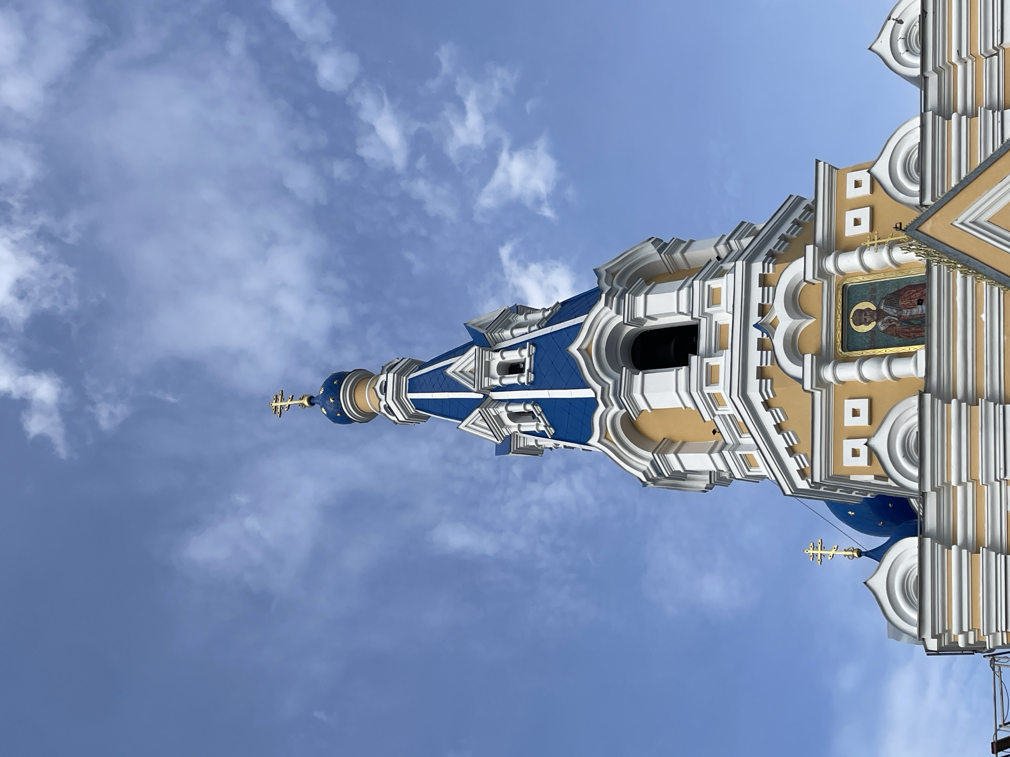 The image shows the exterior of a brightly colored Eastern Orthodox church, likely located in the city of Brest, Belarus. The church features a tall, tapering blue-domed spire topped with a golden cross. The building itself is predominantly yellow with white decorative trim, including arched windows and cornices. Smaller blue domes with golden crosses are visible on lower sections of the building. A mosaic icon of a robed figure with a halo is mounted on the facade. The sky above is a bright blue with scattered white clouds, suggesting daytime. The perspective is from below, looking up at the church's architecture.
