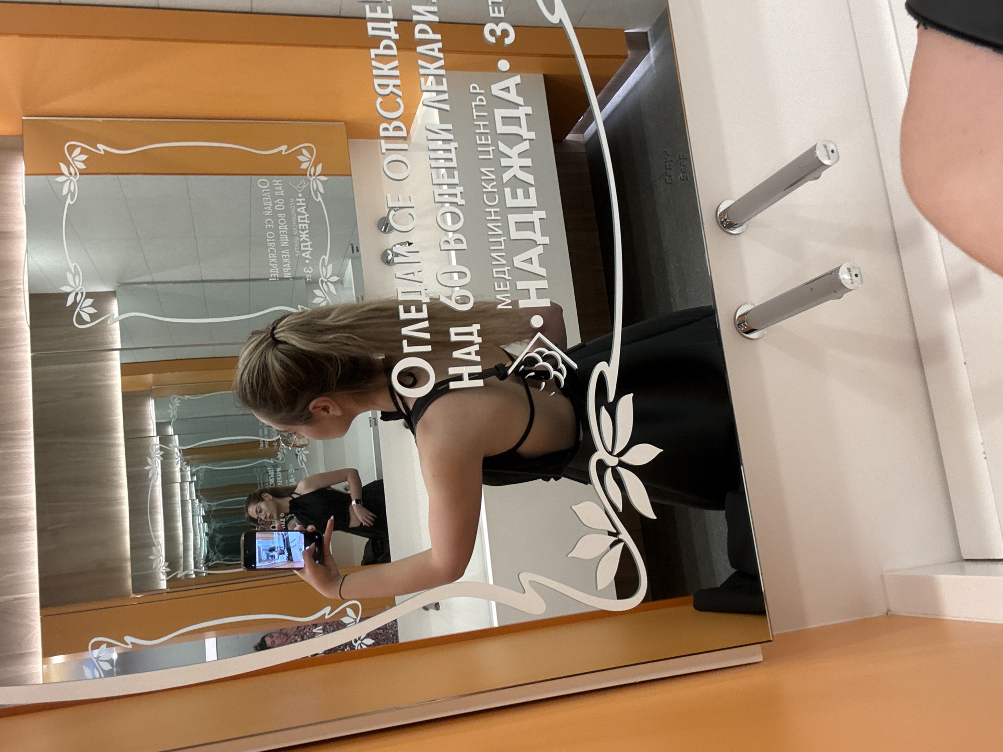 Here is a description of the image:
A woman with blonde hair is taking a mirror selfie in a medical center in Sofia, Bulgaria. The reflection shows her holding a phone, with a picture of herself taking a mirror selfie. The text on the mirror reads: "ОГЛЕДАЙ СЕ ОТВСЯКЪДЕ. НАД 60-ВОДЕЩИ ЛЕКАРИ МЕДИЦИНСКИ ЦЕНТЪР НАДЕЖДА • ЗЕТ. ПАД СЯКЪДЕ! ЛЕКАРИ JA-3 OLVENON CE OLBCUKPE HVU GO BOVEMN VEKVON WEUNΙΩΝΗCKN ПЕНІРЬ ВЕ АДЖЗДАН". The background includes a wall with decorative floral patterns. The center is likely a branch of the Nadezhda Women's Health Clinic.