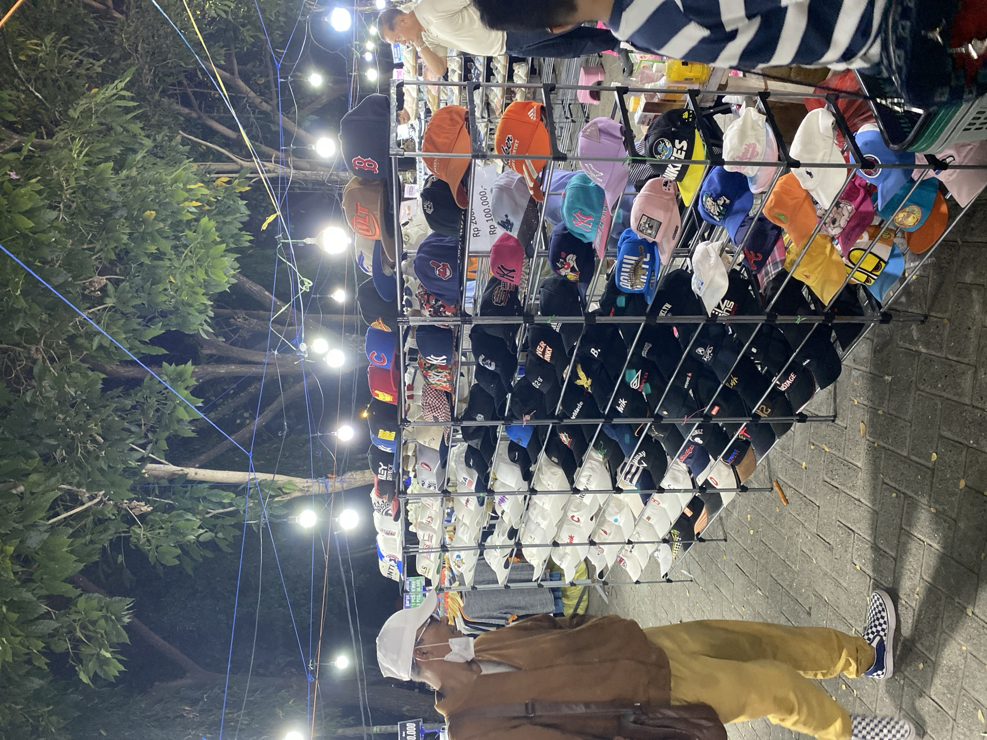 A man wearing a white cap, face mask, brown jacket, and yellow pants walks past a stall filled with various baseball caps at night in Banjarmasin, Indonesia. The stall, illuminated by strings of light bulbs, displays caps neatly arranged on multiple shelves. Some caps feature prominent logos and designs. In the background, another man in a white shirt stands near the stall, and shelves of what appear to be shoes are visible further back. The ground is paved with grey interlocking bricks, and trees with green foliage are present in the background, suggesting an outdoor market setting. The overall scene depicts a bustling night market environment.
