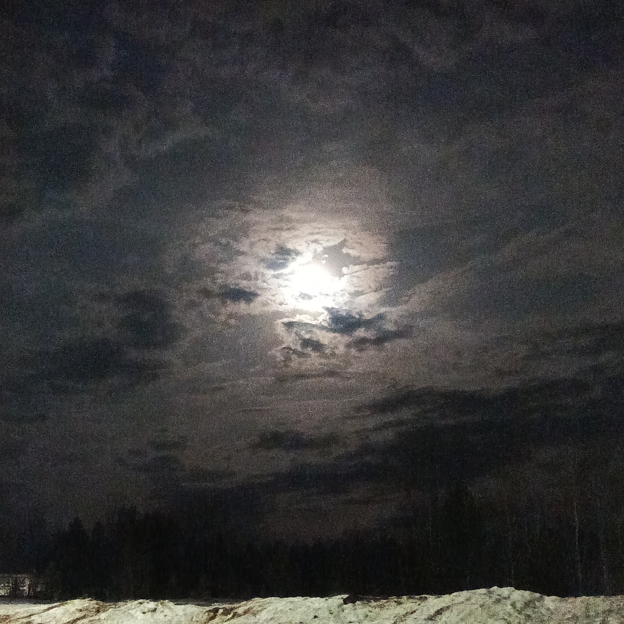 The image depicts a night scene, presumably in a rural or semi-rural area near the city of Марковский, Russia. The sky is filled with dark, heavy clouds partially obscuring the moon, which is the primary light source in the image. The moon's light filters through the clouds, creating a diffused, glowing effect around its center.
In the lower portion of the image, there is a snow-covered foreground with undulating surfaces, likely a snowdrift or a field. Behind the snow, a dark line of trees or forest is visible, forming a stark silhouette against the night sky. The overall impression is one of a cold, possibly desolate winter landscape illuminated by moonlight. No people or animals are visible. A faint, distant structure appears to the left of the forest line.