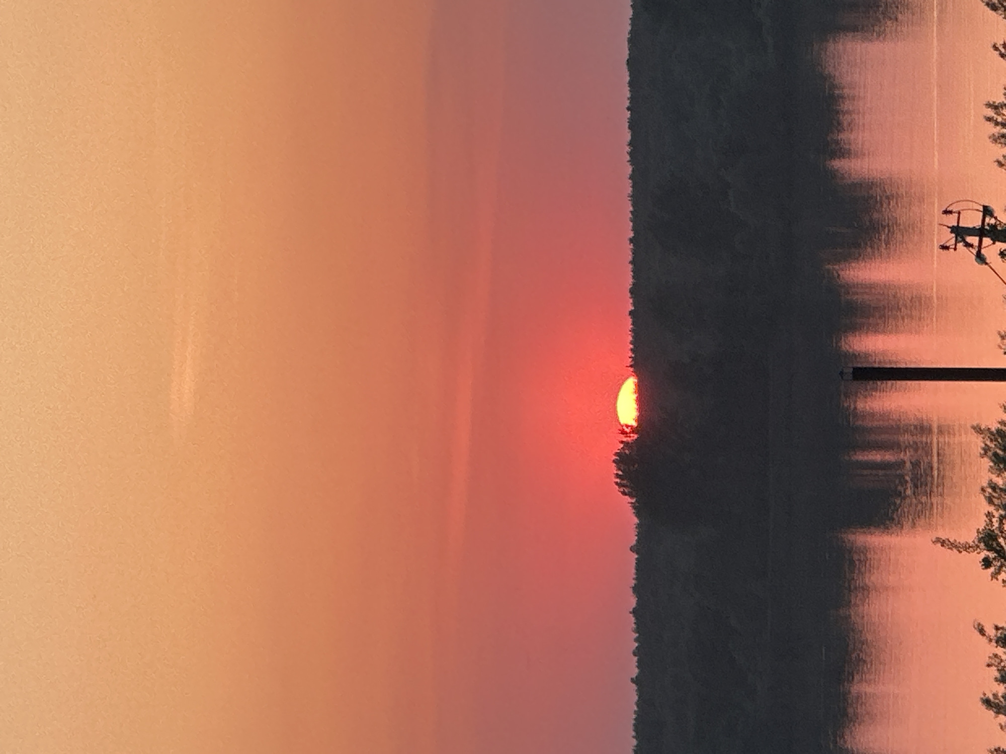 The image depicts a sunset over water, likely a river or lake, with a dark shoreline of trees silhouetted against the sky. A portion of the sun is visible, appearing as a bright red-orange disk partially obscured by the trees. The sky transitions from a lighter orange-peach color near the horizon to a deeper red-orange higher up. On the right side, a utility pole with lines is visible. The water reflects the colors of the sky and the silhouette of the trees. The location is assumed to be in the city of Chaikovsky, Russia.