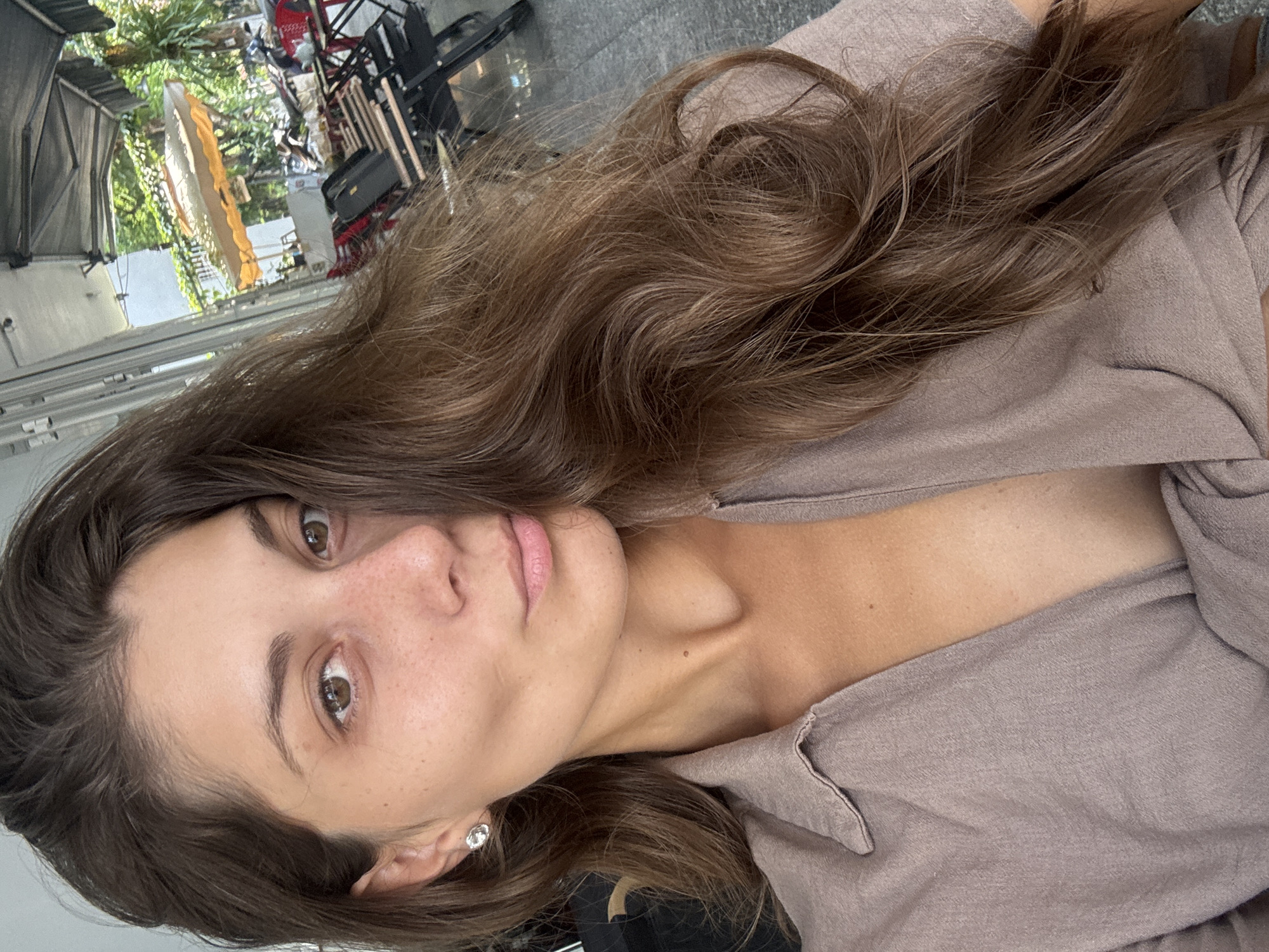 A selfie shot from a slightly low angle, the woman's face is the focal point. She has brown hair, brown eyes, and fair skin with freckles. She's wearing a neutral-colored blouse, and a stud earring is visible in her left ear.
The background shows a blurred outdoor setting, with hints of trees, a white structure that might be a tent or canopy, and equipment that suggests a film or photo shoot location. A red director's chair is visible.
Due to the limited information, I cannot confirm if the location is in Đà Nẵng, Vietnam.