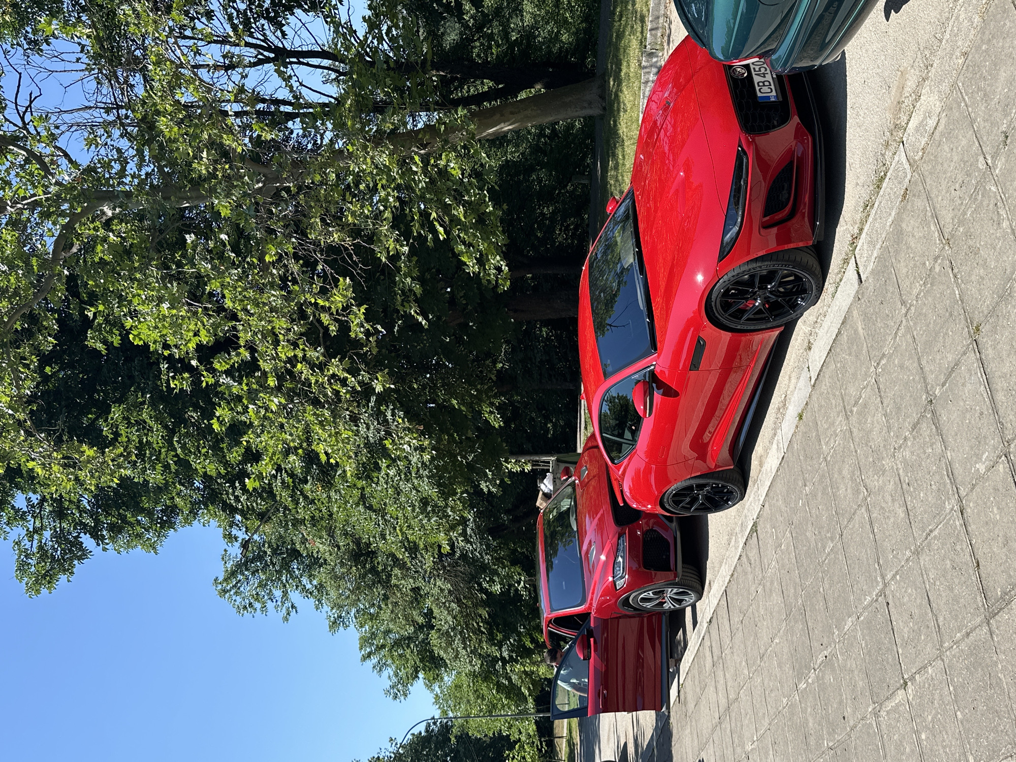 The image captures a sunny day in Veliko Tarnovo, Bulgaria, featuring two vibrant red Jaguar cars parked on a paved walkway next to a grassy area. The foreground is dominated by the sleek, low-profile of a red Jaguar F-Type, its black rims and aggressive front grille prominent. To its left, a red Jaguar F-Pace SUV is parked with its driver's side door open, revealing a person inside. The cars are positioned on a light-colored tiled pavement, with a curb separating it from the road. In the background, lush green trees form a dense canopy, their leaves rustling in the clear blue sky. The overall scene suggests a pleasant outdoor setting, possibly a park or a quiet street in the city. The license plate on the F-Type reads "CB 450".