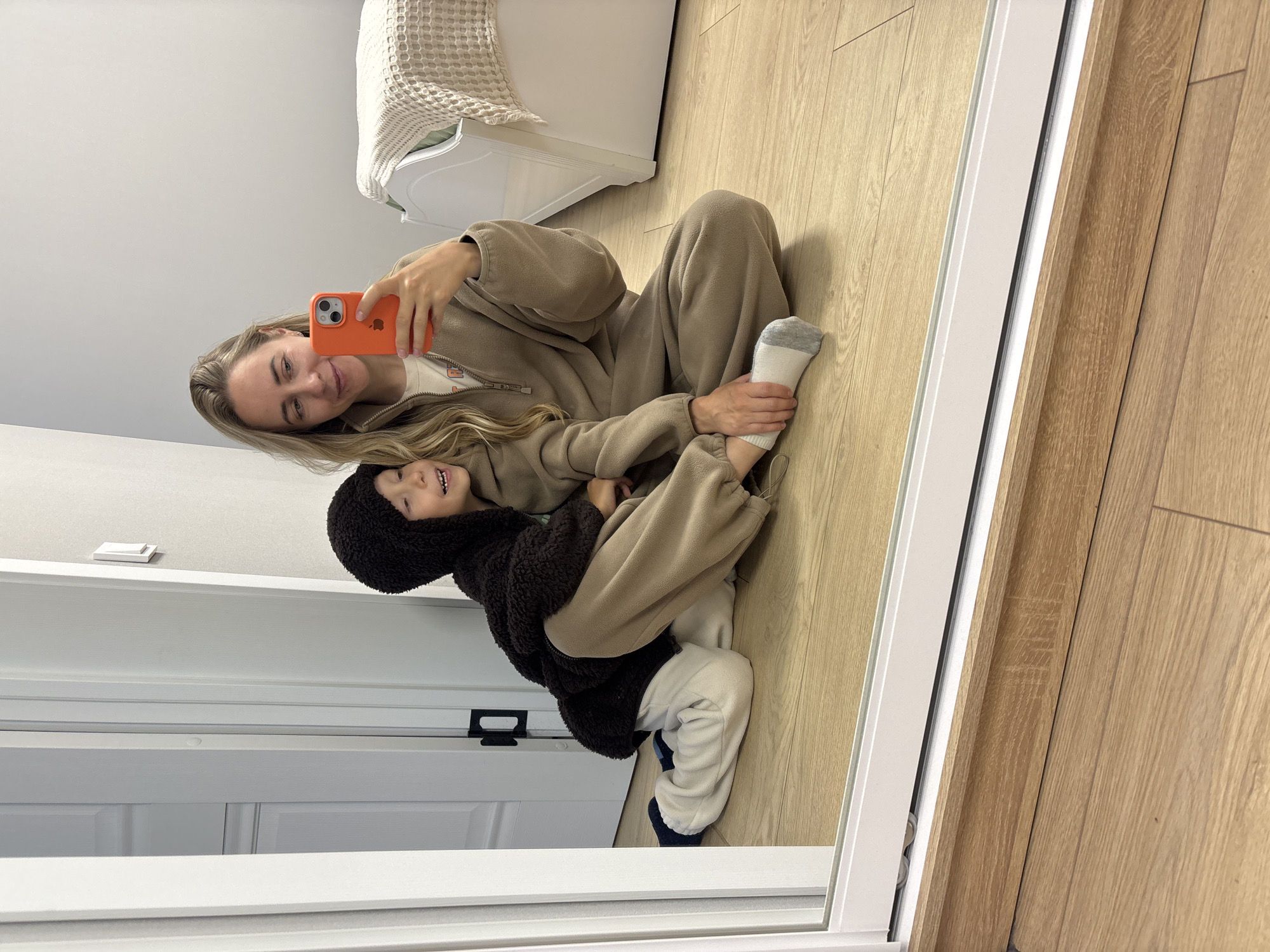 A blonde woman and a young child are sitting on a wooden floor in front of a mirror, taking a selfie. The woman is wearing a beige loungewear set and holding an orange phone. The child is wearing a dark fuzzy hooded jacket and white pants. They are both smiling. The background shows a white door and a bed with a beige blanket. The room appears to be a bedroom. There is no information in the image to suggest the location is the city of Chaikovsky, Russia.