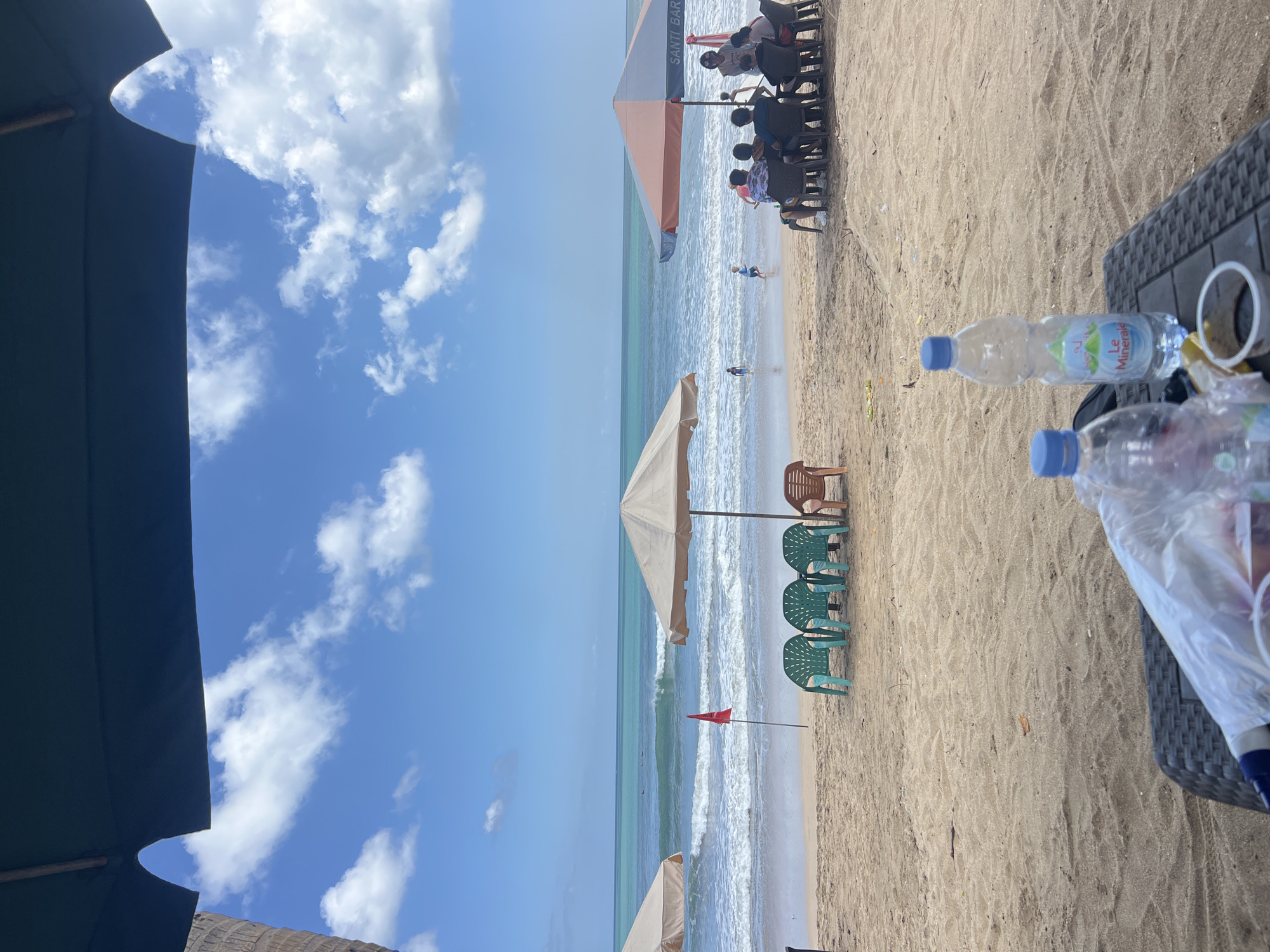 This is a photograph taken at a beach in Kuta, Indonesia, likely on a sunny day with a few clouds. The foreground features a plastic table with two bottles of water, one labeled "Le Minerale," and a small white cup. Beyond the table, a sandy beach is visible with several rows of beach chairs under large umbrellas. On the left, there are two beige umbrellas, and on the right, a larger umbrella with orange and blue panels displays the text "SANTI BAR." A group of people are seated at tables under the right umbrella, while three green plastic chairs and one brown plastic chair are unoccupied in the middle of the beach. A red flag is planted in the sand near the water's edge. The ocean in the background has moderate waves, and a few individuals, possibly children, are visible in the shallow water. The overall scene depicts a relaxed beach environment.
