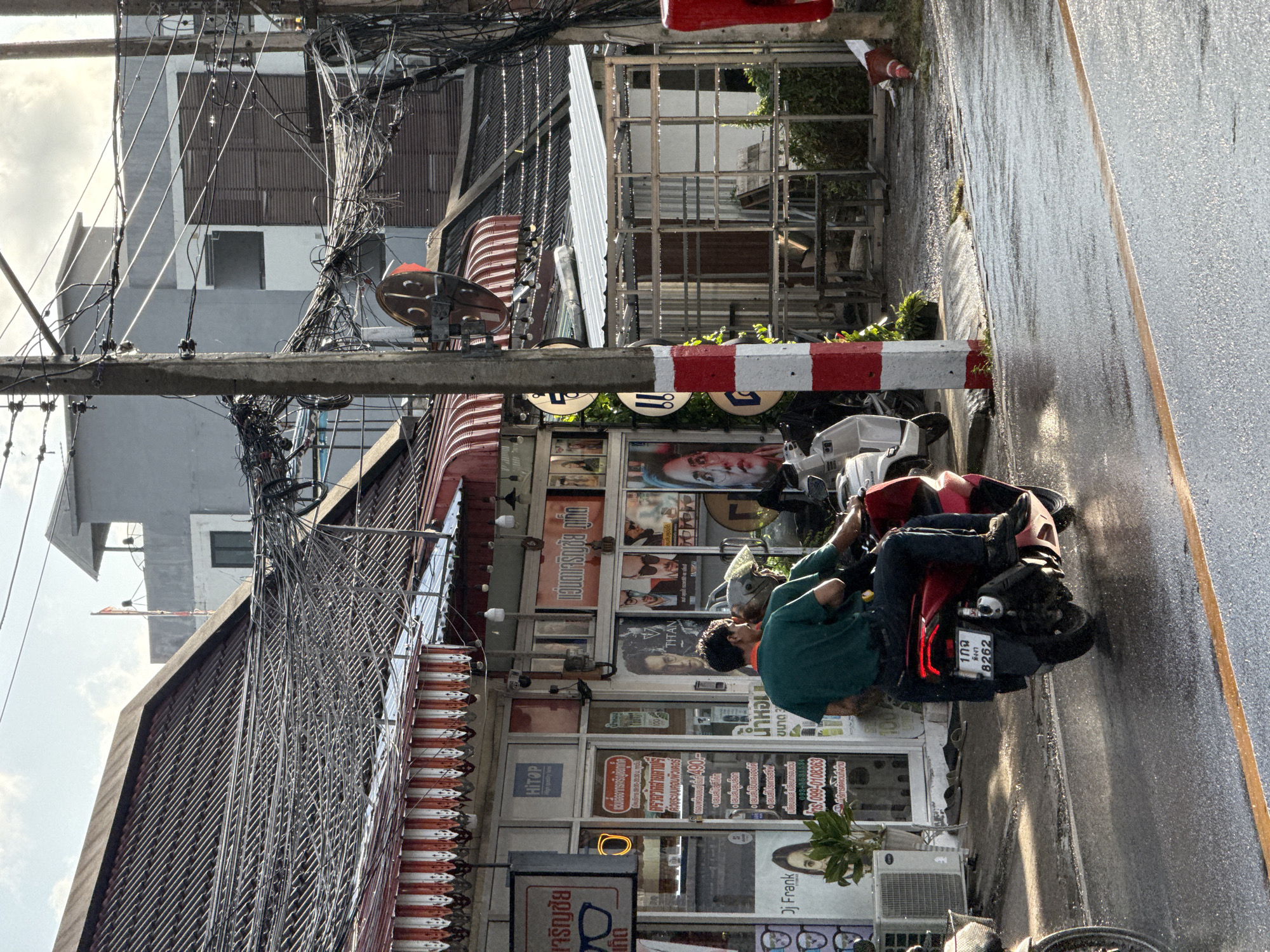 This is a medium shot of a street scene in Wichit, Thailand, taken on a cloudy day. The foreground is dominated by a wet asphalt road with a yellow line on the right side, indicating a lane. In the middle ground, a red scooter with two occupants is moving away from the viewer. The driver is wearing a green t-shirt and dark pants, and the passenger is wearing a helmet and a darker shirt. The scooter has a license plate with Thai characters and the number "8262". To the left of the scooter, a white scooter is parked.
The background features a row of shops with tiled roofs and awnings. Some of the shop windows display posters and signs with text in Thai. A prominent concrete utility pole with numerous tangled wires runs vertically through the center of the image. The pole is painted with alternating red and white stripes. Behind the shops, there are more buildings and tangled overhead wires, characteristic of urban areas in Thailand. The overall impression is of a busy street with dense infrastructure.