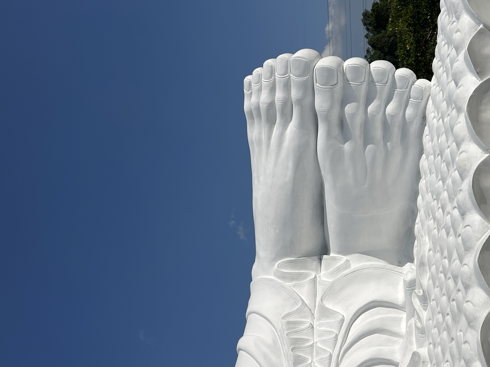 The image shows a close-up view of the feet and lower legs of a large, white, reclining statue, likely a Buddha. The statue is made of a smooth, matte material and is detailed with folds that suggest drapery or clothing. The feet are prominently displayed, with clearly defined toes and toenails. The statue rests on a textured surface that appears to be part of its base, possibly depicting lotus petals or waves.
In the background, the sky is a clear, deep blue with a few wispy white clouds. To the right, a line of lush green trees and foliage is visible, bordering a bright, slightly hazy horizon. A faint power line can also be seen against the sky. The overall setting appears to be outdoors, under bright daylight.
This statue is located in the city of Pupuan, Indonesia. The statue is a significant landmark and religious icon in the region.