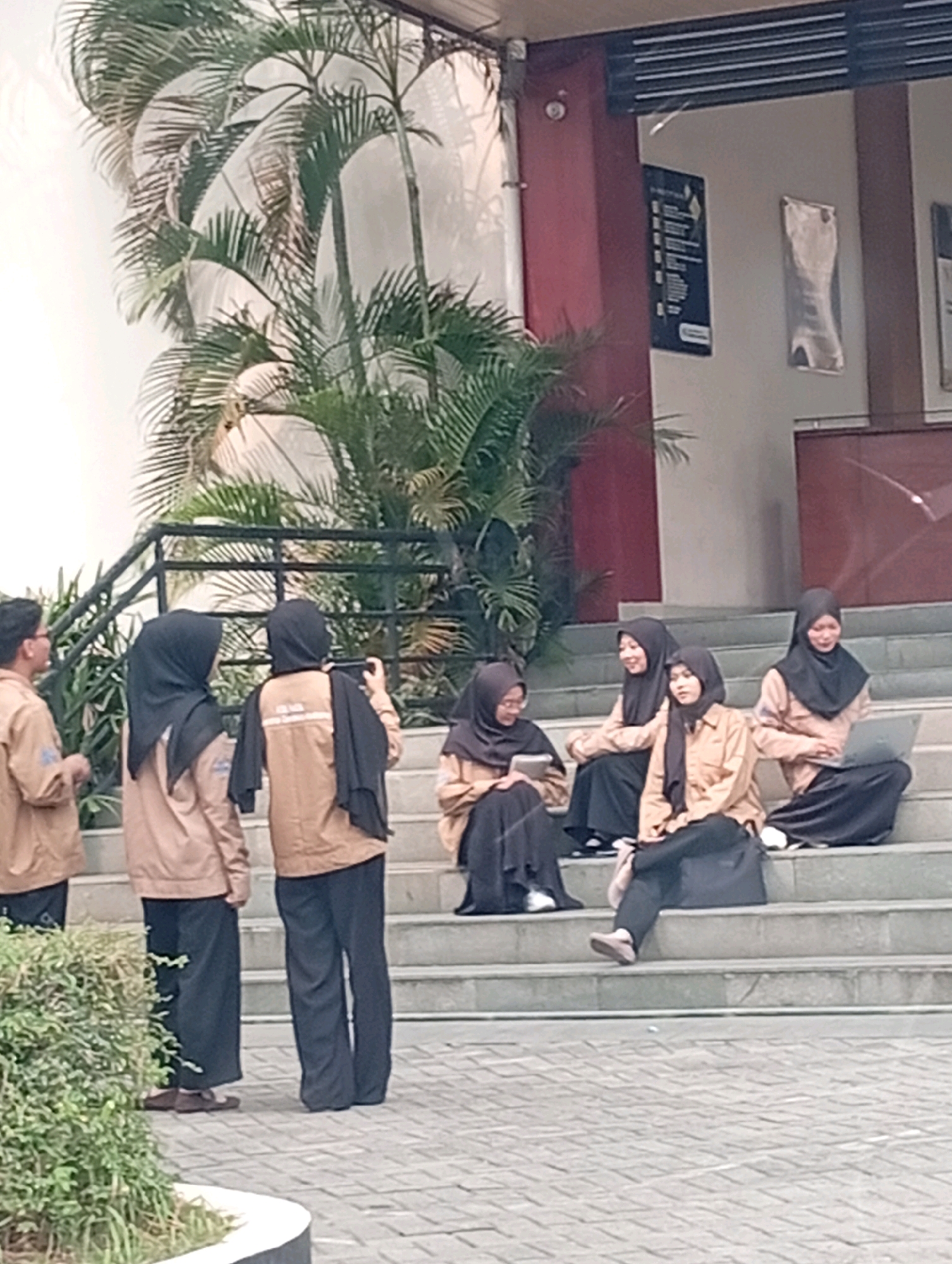 A group of five young women, dressed in matching light brown jackets and dark hijabs, are seated and standing on a set of outdoor concrete stairs. The women are positioned in two groupings: three are seated on the stairs, and two are standing further up. One seated woman on the far right is looking at a laptop. Another seated woman to her left appears to be looking down. The third seated woman, in the middle, is looking forward. The two standing women are facing away from the camera. The woman on the left is looking to her side, and the woman on the right is holding a phone. In the background, there are large palm fronds from a plant, and the exterior wall of a building, painted in a dark reddish-brown and a lighter beige color. The stairs appear to be part of an outdoor walkway or entrance to a building. The scene is likely a public outdoor space in a city like Kelapa Dua, Indonesia, given the presence of palm trees and the architectural style.