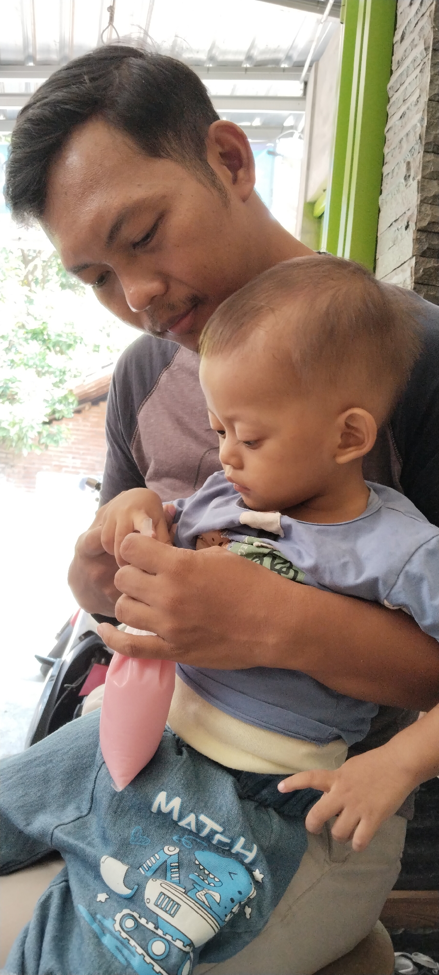 In Peganjaran, Indonesia, a man and a young child are seated. The man, with dark hair and a mustache, is looking down at the child, who is held in his arms. The child, wearing a light blue shirt with a dinosaur graphic on the pants, is also looking down, appearing to interact with a small pink pouch held by the man's hands. The background suggests an outdoor or semi-outdoor setting, with glimpses of greenery and possibly a building structure. The overall scene depicts a moment of shared activity and attention between the man and the child.