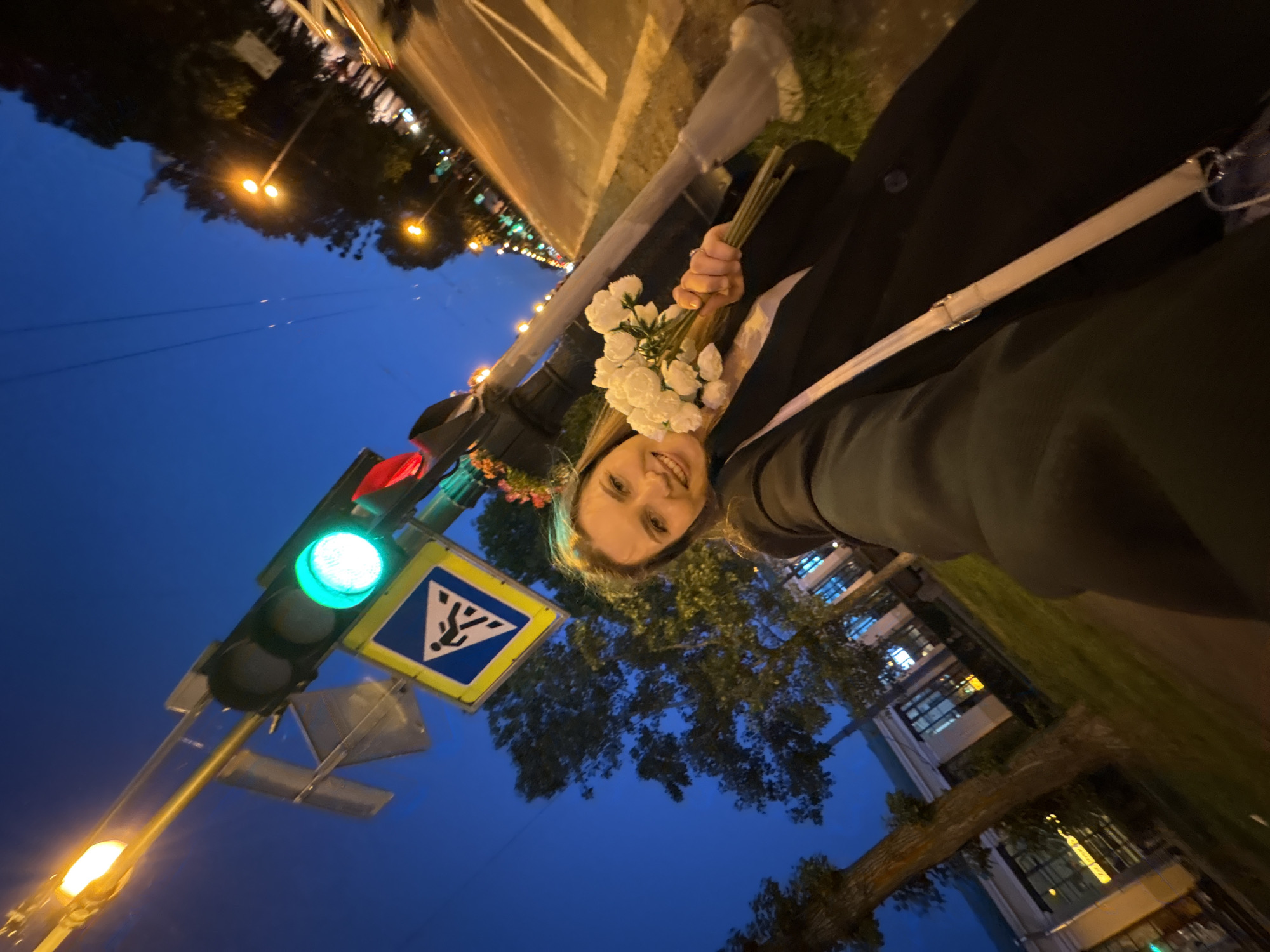A selfie shows a smiling young Caucasian woman holding a bouquet of white artificial flowers while standing at a pedestrian crossing at night. She is wearing a dark coat. In the background, a traffic light shows a green signal, and a pedestrian crossing sign is visible. Streetlights and buildings line a road, indicating an urban environment. The dark blue sky suggests it is evening or early night. The image appears to be taken on a street in Saint Petersburg, Russia, likely in a well-lit area with pedestrian infrastructure.