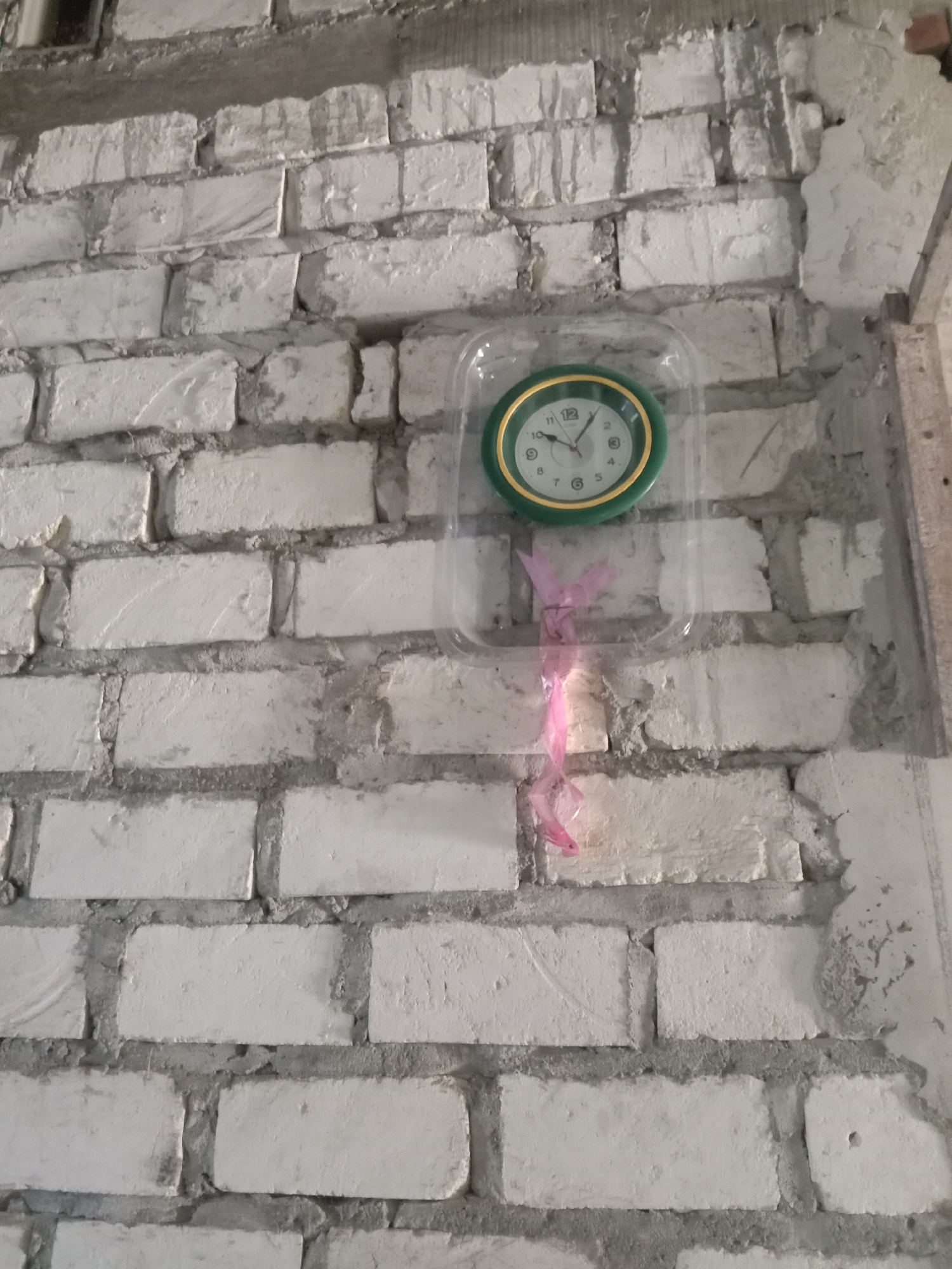 A clock is mounted on a rough white brick wall. The clock has a green bezel and a white face with black numbers and hands. A pink ribbon is tied to the bottom of the clock and hangs down. The wall appears to be unfinished or in disrepair. There are no people or animals visible in the image. The scene is likely an interior or exterior wall in a construction or renovation area in Stiaji, Indonesia.