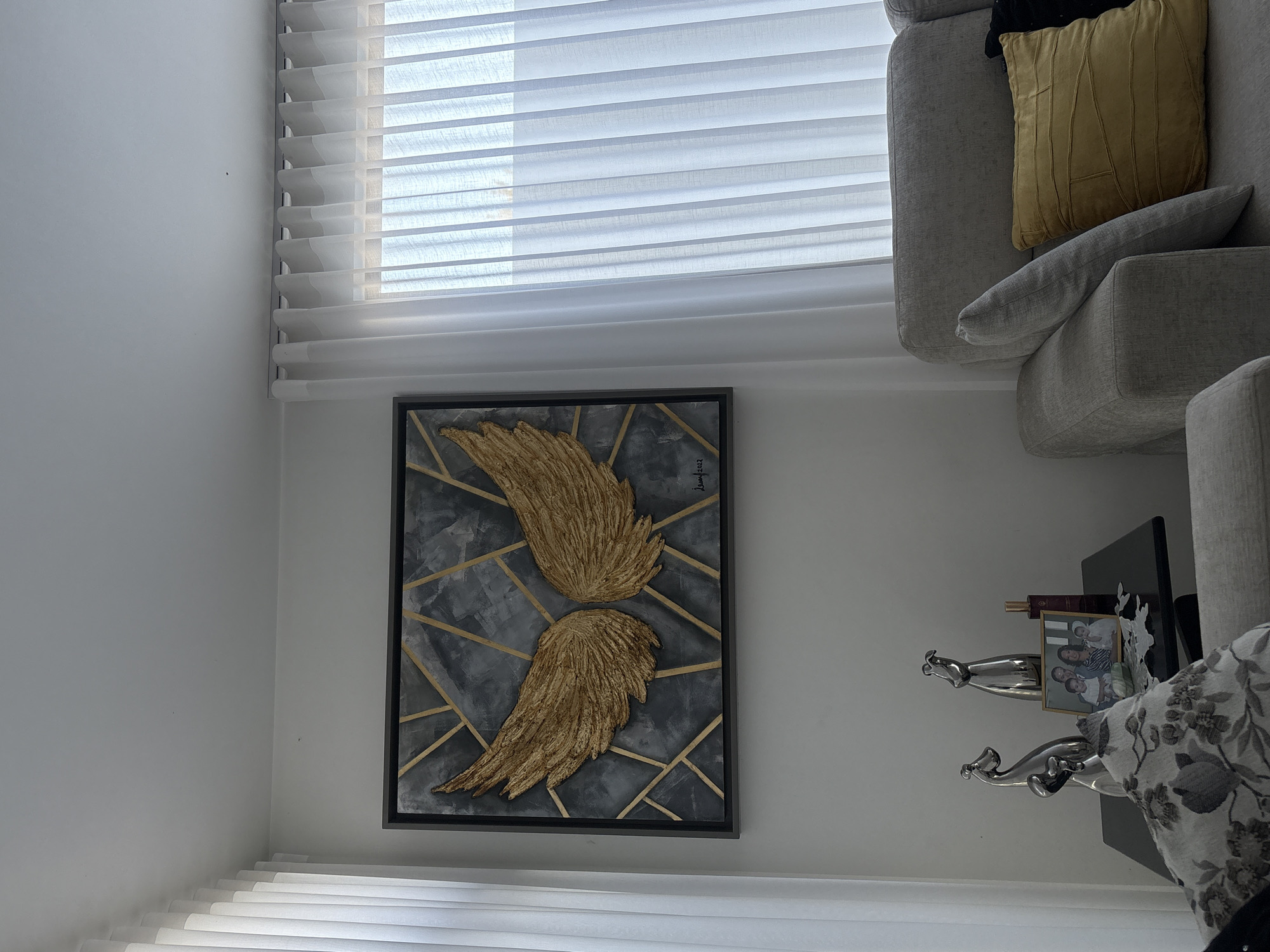 The image shows an interior corner of a room with a partially open window covered by horizontal blinds at the top. Below the window is a framed piece of art depicting two golden wings against a grey background with gold geometric lines. To the right of the artwork, a light gray sofa is partially visible with a gold colored pillow. Beside the sofa is a small black table with two silver jaguar statues, a framed photograph, and other items on it. The walls are painted white. No people are visible. The location is unspecified, not San Bernardino Tlaxcalancingo.