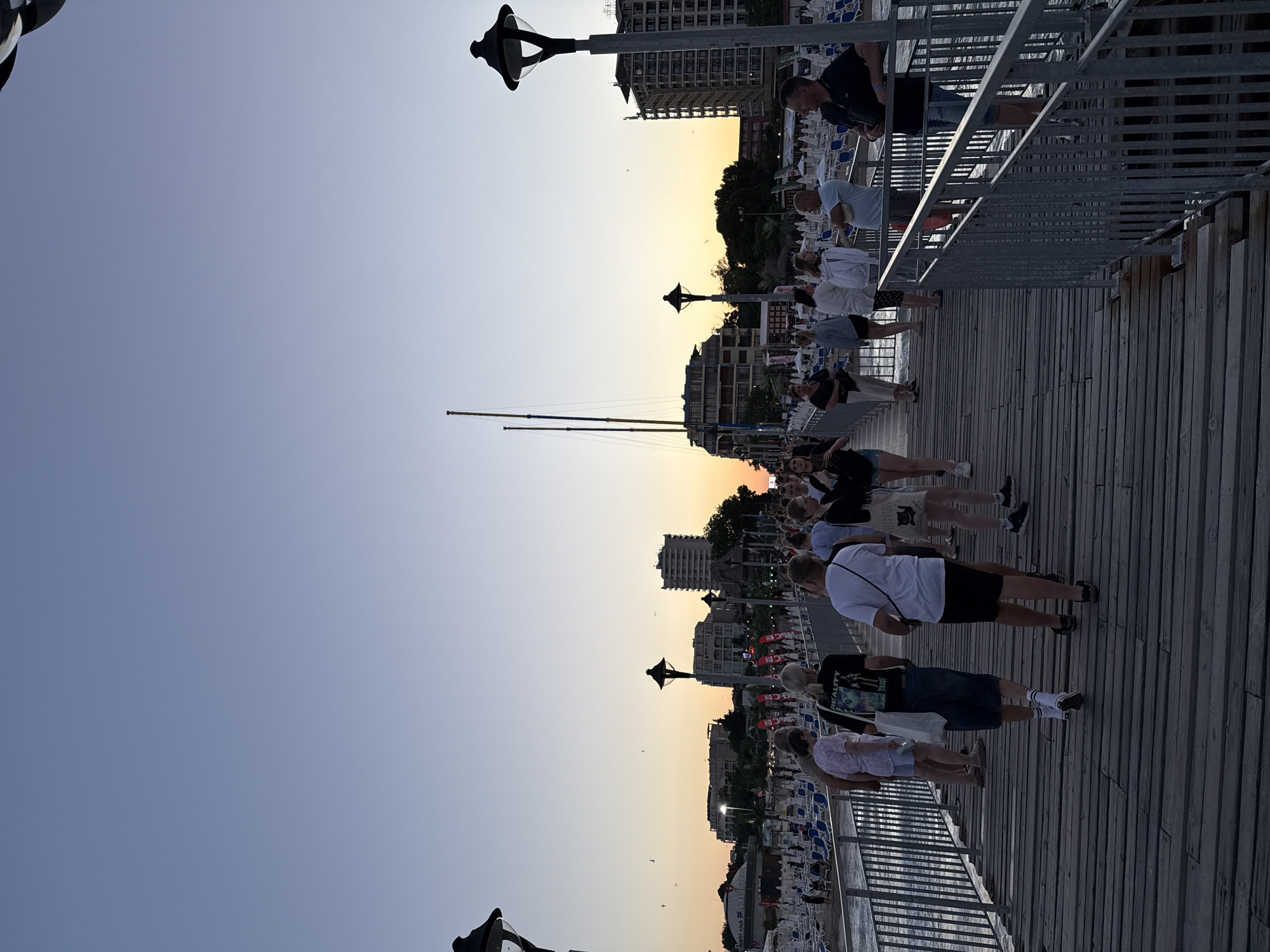 This image depicts a view from a wooden pier in Nesebar, Bulgaria, looking towards a beach and a cityscape at sunset. The sky is a clear gradient of pale blue to orange, indicating the time of day.
Several people are walking along the pier, their backs mostly to the camera. They are dressed in casual summer clothing. On the left side of the pier, there are rows of empty beach chairs and umbrellas, suggesting a public beach area. In the background, buildings of varying heights, including hotels and what appears to be a nightclub with a distinctive circular roof, are visible. Two tall, slender masts, possibly from a sailboat, rise above the buildings.
Streetlights with ornate black lanterns are positioned along the pier, some of which are lit. A metal railing runs along the edge of the pier. The wooden planks of the pier are visible in the foreground, showing signs of wear. The sea is visible to the left of the pier, with gentle waves. Several birds are flying in the sky. The overall atmosphere is one of a relaxed evening scene at a seaside resort.