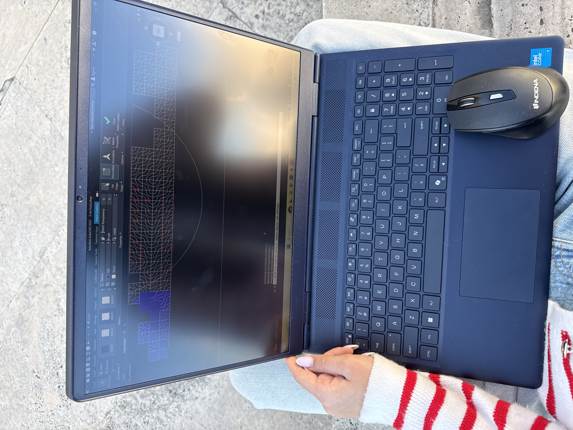 A person sits outside with a laptop on their lap. The laptop screen displays what appears to be Autodesk AutoCAD 2022, with a 3-D design and various menus visible. A black Indena wireless mouse sits on the laptop’s right side. The laptop is dark blue, and an Intel Core i7 sticker is attached. The person wears light-wash jeans and a white sweater with red stripes. The location is Yerevan, Armenia, but the specific area is not visible.