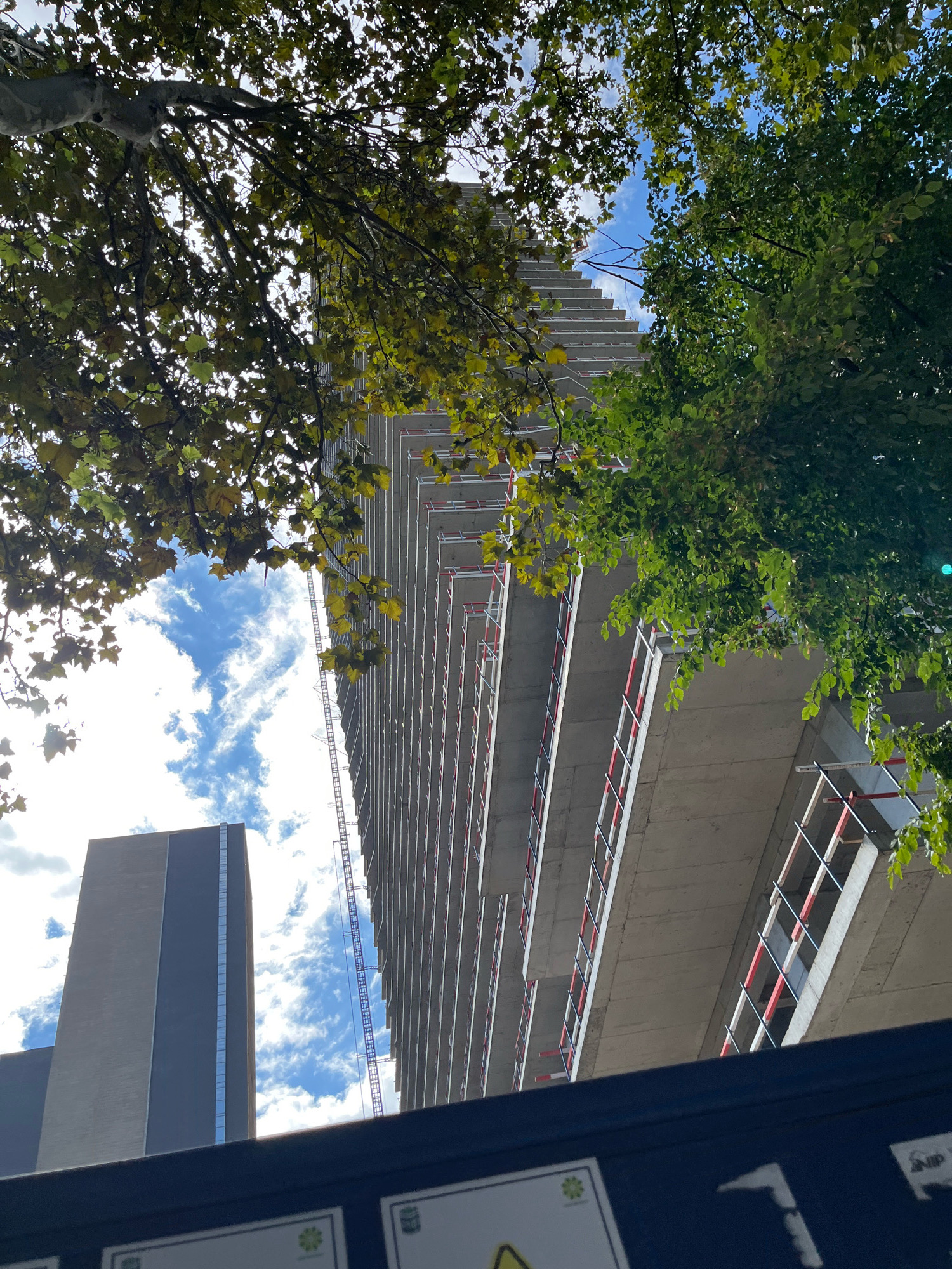 This is a low-angle, upward-facing photograph of a construction site with mature trees framing the view. A tall, unfinished concrete building with multiple floors is the central subject, its exterior showcasing exposed concrete slabs and scaffolding with red and white railings. To the left, a dark, rectangular building with a smooth facade and a vertical strip of windows stands partially obscured by trees. The sky above is bright blue with scattered white clouds. The photograph appears to have been taken on a sunny day, with sunlight filtering through the dense green foliage of the trees, casting dappled shadows. The overall impression is one of urban development contrasted with natural elements.