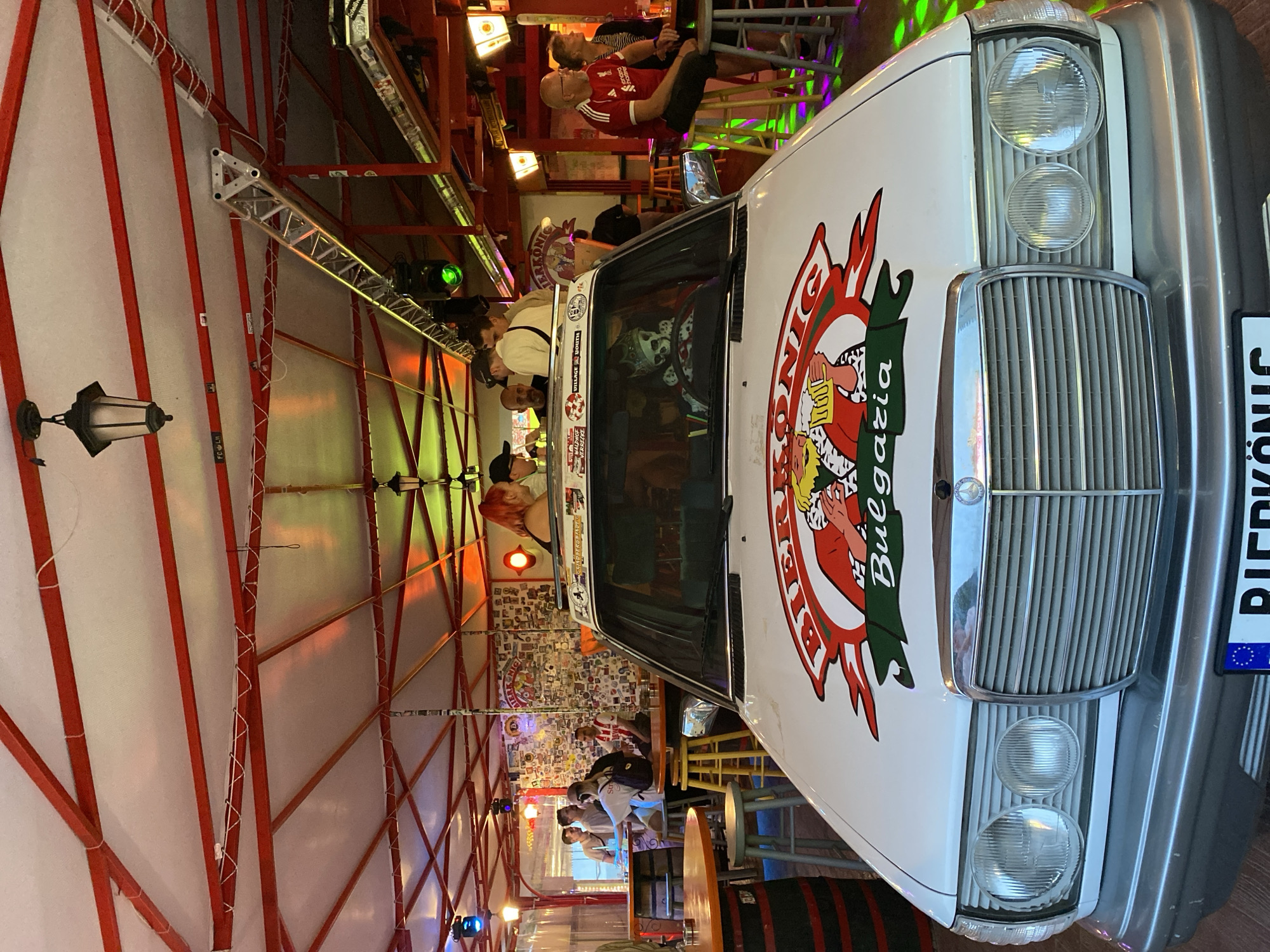 The image captures an indoor or covered outdoor bar scene, specifically identified as "Bierkönig Bulgaria" located in Varna, Bulgaria.
The central subject is a white Mercedes-Benz W124 series sedan, prominently parked with its hood facing the viewer. The hood features a large, colorful decal depicting a crowned figure holding a beer mug, with "BIERKÖNIG" above and "Bulgaria" below. A skeleton figure wearing a crown is positioned in the driver's seat. The car's license plate, partially visible, reads "BIERKÖNIG" with the blue Euroband displaying "BG" for Bulgaria. Various stickers, including "WILDPAGE RECORDS" and "VILLAGE YOUTH," are affixed to the roof rack and the top edge of the windshield.
The environment is a lively bar or beer hall setting, characterized by a white canvas roof supported by a visible framework of red metal beams and trusses. Overhead, traditional-style lanterns and modern spotlights (emitting green, blue, and orange light) illuminate the space, casting colorful reflections. White string lights are also strung along the red beams.
Multiple groups of patrons are visible throughout the venue. On the left, several individuals are gathered around wooden barrel-style tables and high-top tables with various stools (yellow, blue). They are engaged in conversations, some holding drinks. Further right, near the car's passenger side, a man in a red Liverpool FC shirt and an older woman are seated at a high-top table with a drink. In the background, a wall is extensively covered with numerous stickers, resembling a collage, and a neon sign (partially visible) bears the "Bierkönig" name. The overall atmosphere suggests a social gathering point, likely in the evening.