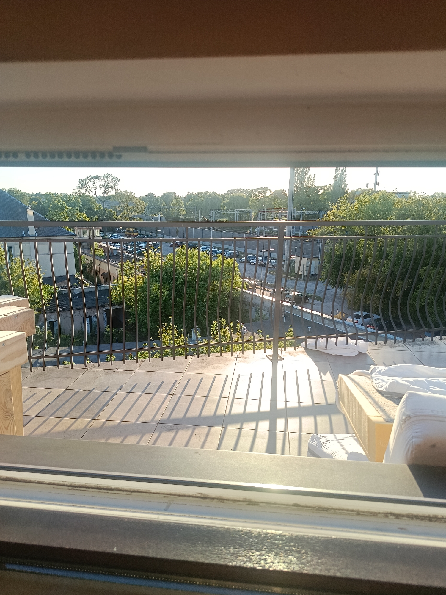 The image shows a view from a balcony overlooking a street and surrounding greenery in Grodzisk Mazowiecki, Poland. The foreground features a tiled floor with shadows cast by a metal railing, indicating it is daytime. A partially assembled bed or mattress is visible on the right side of the balcony. Beyond the railing, a parking lot filled with cars and a yellow van is situated next to a low-rise building and overgrown vegetation. Further in the background, trees and some industrial structures are visible under a bright, possibly sunset sky. The scene appears to be an urban or suburban environment with a mix of residential and commercial elements.