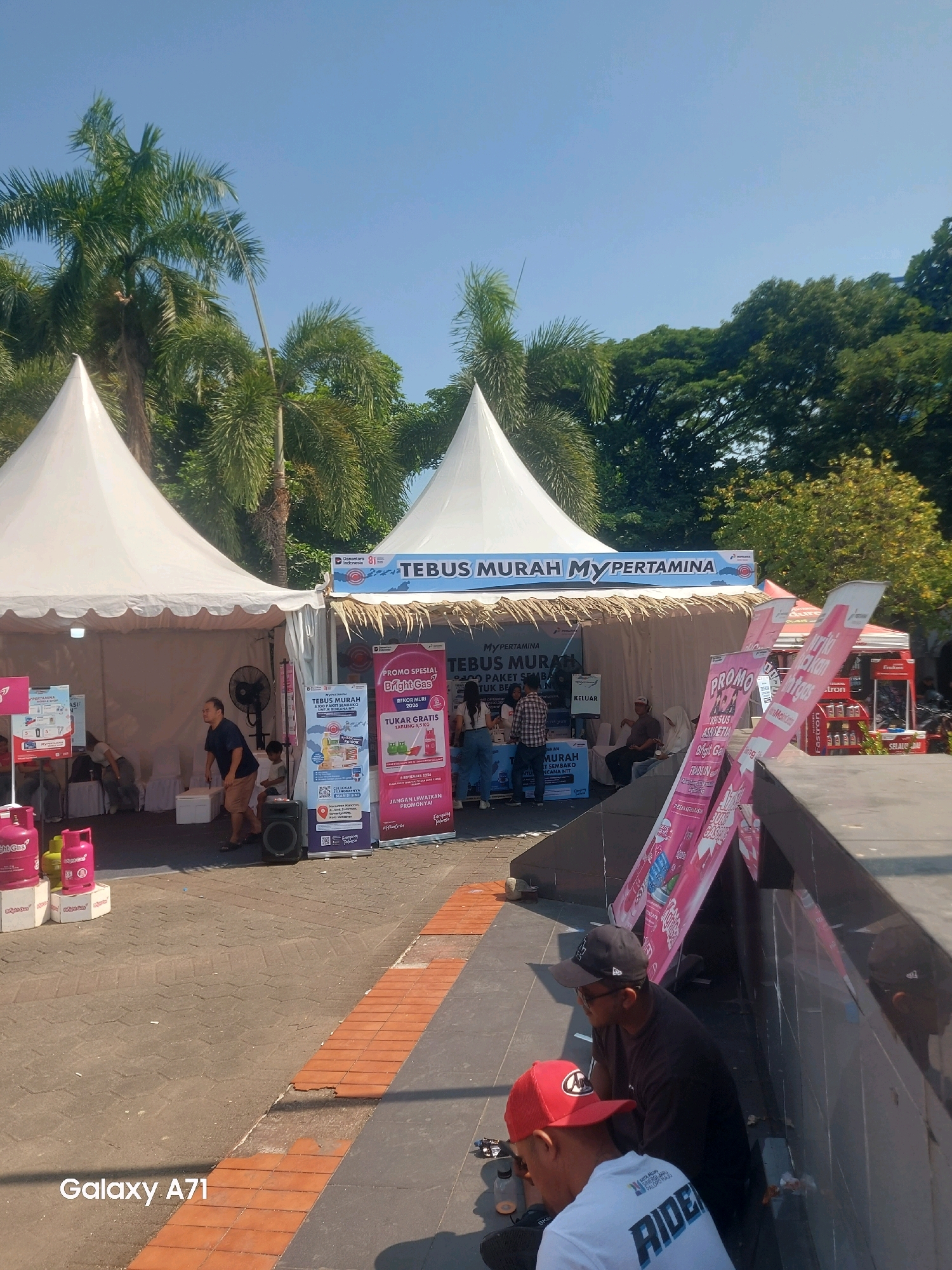The image depicts an outdoor promotional event under a clear blue sky, likely in the city of Makassar, Indonesia, given the "Galaxy A71" watermark. Two white tents, possibly part of a fair or exhibition, are set up, with palm trees and lush green foliage visible in the background. A large banner above one of the tents reads "TEBUS MURAH MY PERTAMINA," suggesting a promotion related to the Pertamina energy company.
In front of the tents, there are several promotional displays and some people interacting with them. Pink gas cylinders are visible near the left tent. A man in a dark blue t-shirt and brown shorts is standing near a fan and a speaker. A young boy is also in the vicinity. On the right side, several pink banners are displayed prominently, advertising "PROMO SPESIAL BRIGHT GAS".
Further to the right, two individuals are seated, with one wearing a red baseball cap and a white t-shirt with the word "RIDER" printed on it, and the other wearing a black t-shirt and a black baseball cap. They appear to be taking a break from the event. The ground is paved with interlocking bricks and some sections with orange tiles, indicating an urban outdoor space. The overall scene is lively, with promotional activities and people congregating under the sunny conditions.