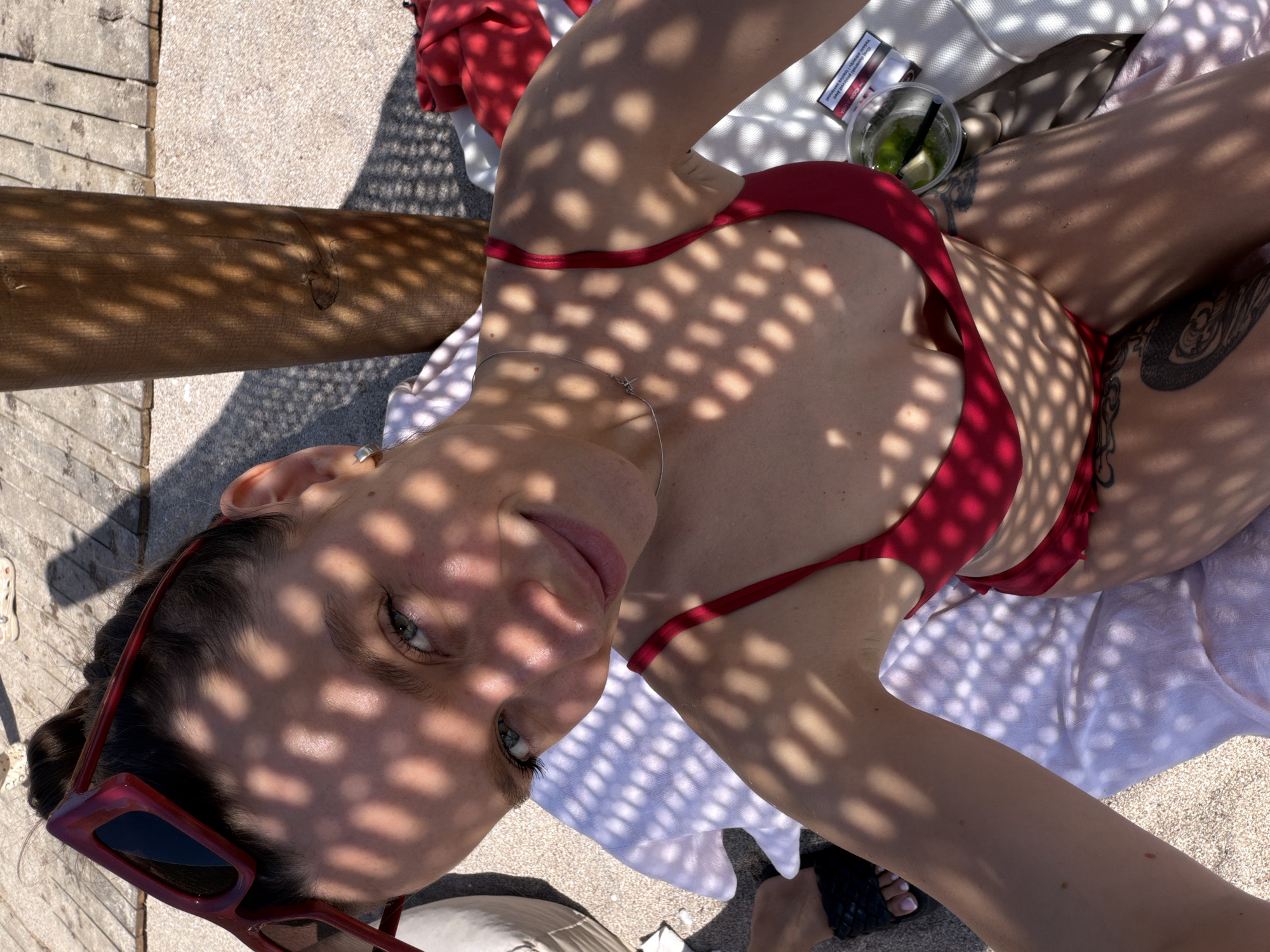 A woman in a red bikini is lying on a lounge chair on a beach in Sutomore, Montenegro. She is looking at the camera and smiling. There are shadows on her face and body from a sunshade. A drink with lime and ice is on the lounge chair next to her. There is also a pack of cigarettes. The background is blurred, but it appears to be sand and water.