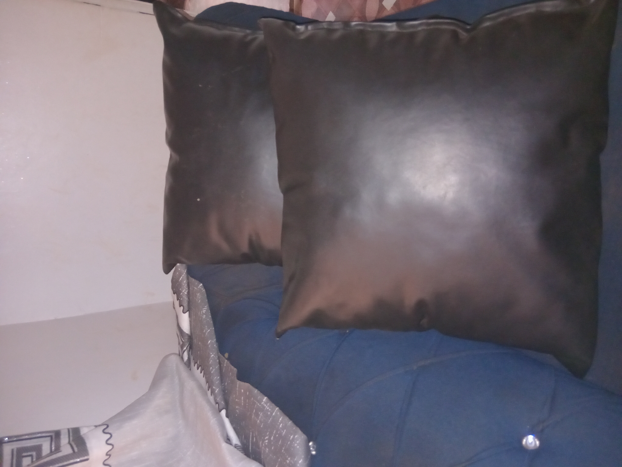 The image shows two dark, possibly faux leather, pillows on a blue tufted sofa. A white and grey patterned decorative item (possibly a pillow or blanket) is visible in the lower left corner. The background is a plain, light-colored wall. There is no visible information in the image that confirms its location is Shagamu, Nigeria, or any information about people, subjects, or actions.