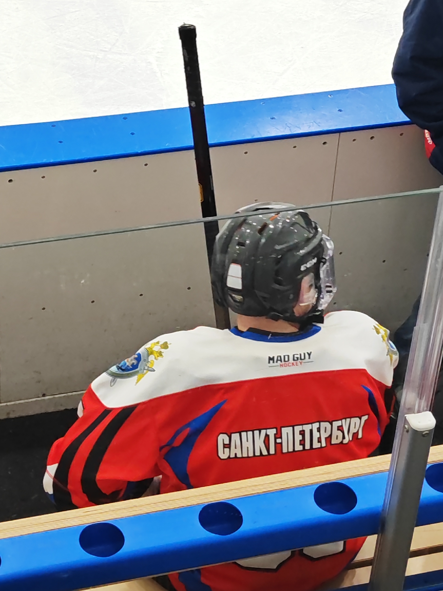 A hockey player wearing a red, white, and black jersey with "САНКТ-ПЕТЕРБУРГ" (Saint Petersburg) and "MAD GUY - HOCKEY" written on it sits on a blue bench in the player box at an ice rink. The ice rink's boards are visible above the bench, with a hockey stick leaning against the boards. A glass barrier separates the bench from the ice rink. Part of another person in a dark blue jacket is visible on the right.