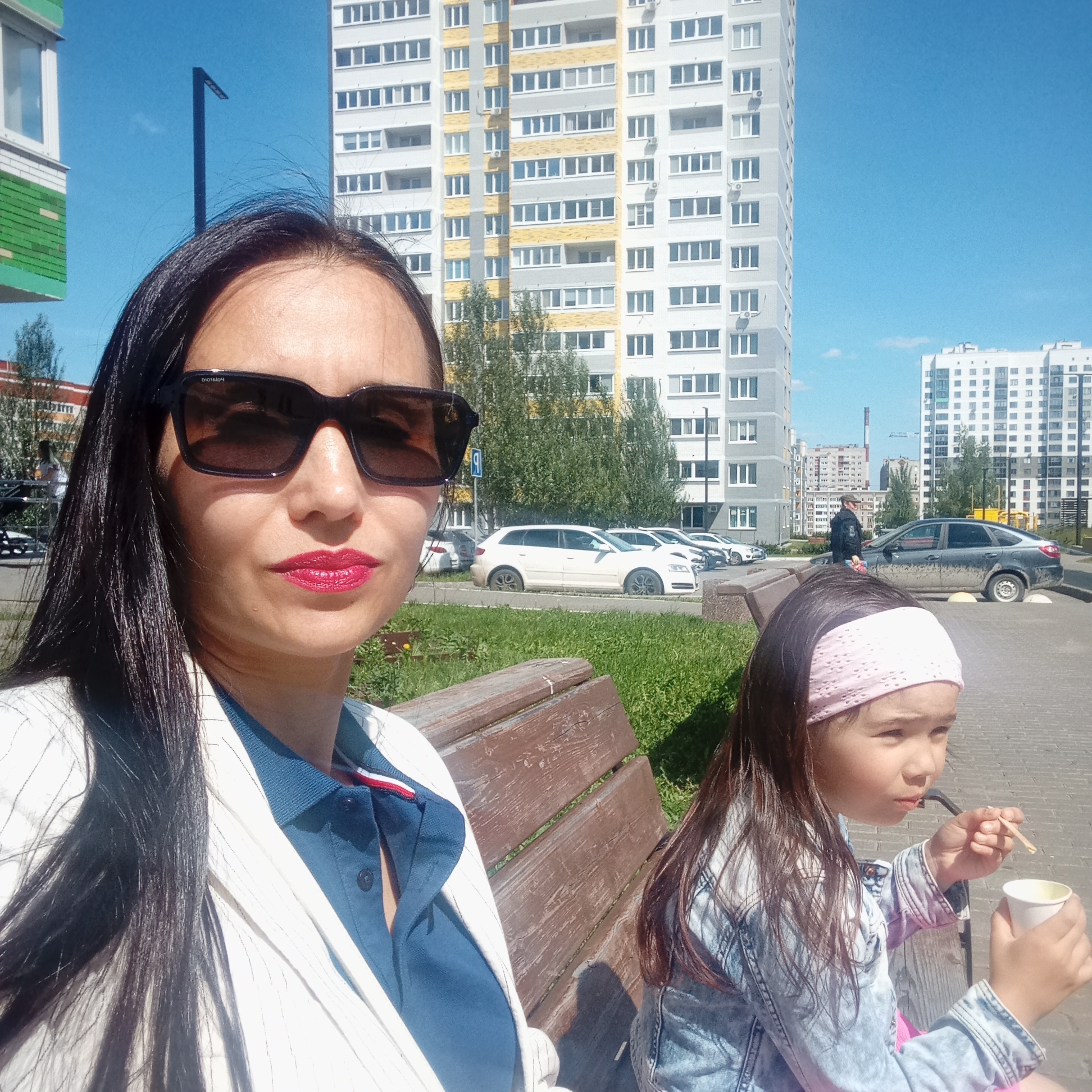 An adult female and a young girl are seated on a wooden bench outdoors in an urban environment, consistent with the city of Izhevsk, Russia, on a clear, sunny day.
The adult female, positioned in the foreground on the left, has long, dark hair and is wearing dark rectangular sunglasses (identifiable as "Polaroid"). She has bright red lipstick and is dressed in a white pinstripe blazer over a dark blue polo shirt featuring a red, white, and blue stripe detail on the collar. She looks directly towards the camera with a neutral expression.
To her right, seated on the same bench, is a young girl with dark hair, wearing a light pink knitted headband and a light blue denim jacket. She holds a white paper cup in her left hand and a thin wooden stick in her right hand, which she brings towards her mouth, indicating consumption. Her gaze is directed to the right, away from the camera.
The immediate surroundings include green grass to the left of the bench and a grey paved area extending into the mid-ground. Parked cars are visible on the paved area, including a white Audi A3 Sportback and a dark grey Lada Granta sedan with noticeable dirt on its lower body. A blue parking sign is also visible. In the background, multiple tall, multi-story residential apartment buildings dominate the skyline under a clear blue sky. One prominent building features a light grey facade with yellow sections, while another is predominantly white. Deciduous trees with green foliage are scattered between the road and the buildings. A tall, slender black street light is visible to the upper left, and a distant industrial chimney can be seen on the horizon to the right. A male figure in dark attire is walking in the background near the parked cars.