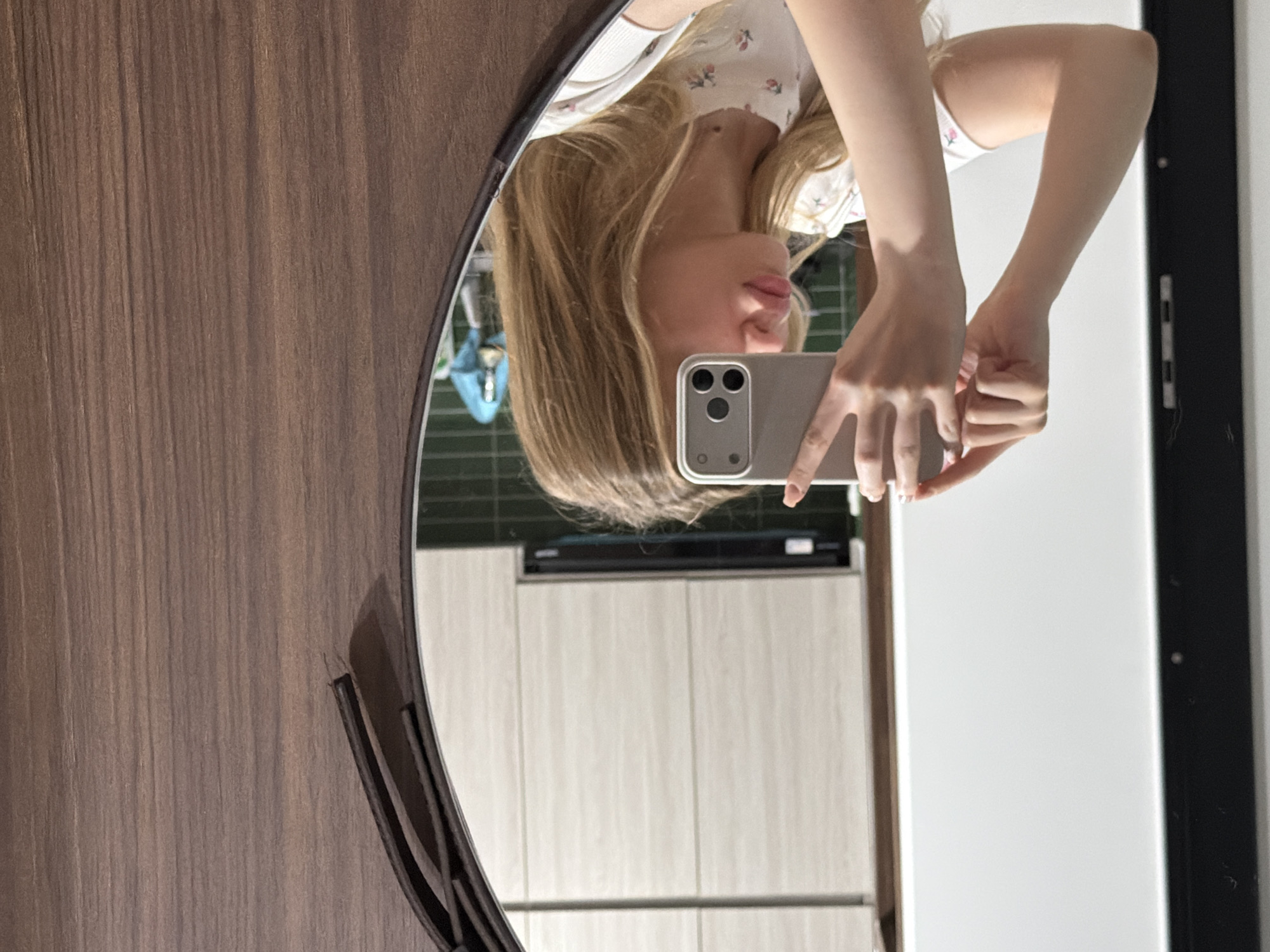A person takes a selfie in a mirror. The person has long, blond hair and is wearing a floral-print top. The phone is gray. The background appears to be a kitchen. The location is not discernible from the image.