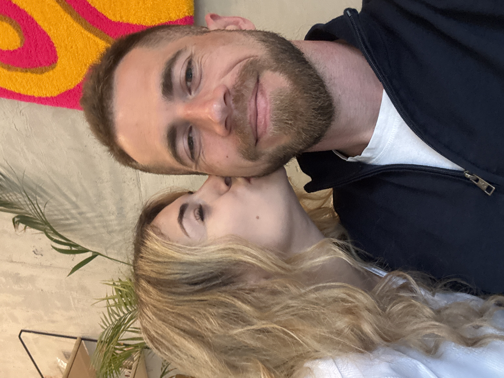 A man with short brown hair and a beard, wearing a black zip-up jacket over a white shirt, is being kissed on the cheek by a woman with long, wavy blonde hair. The woman is wearing a white top. They are indoors, with a wall and some greenery visible in the background. A colorful wall decoration is visible above the man. The location is likely in Budva, Montenegro. No further details about the location are discernible.