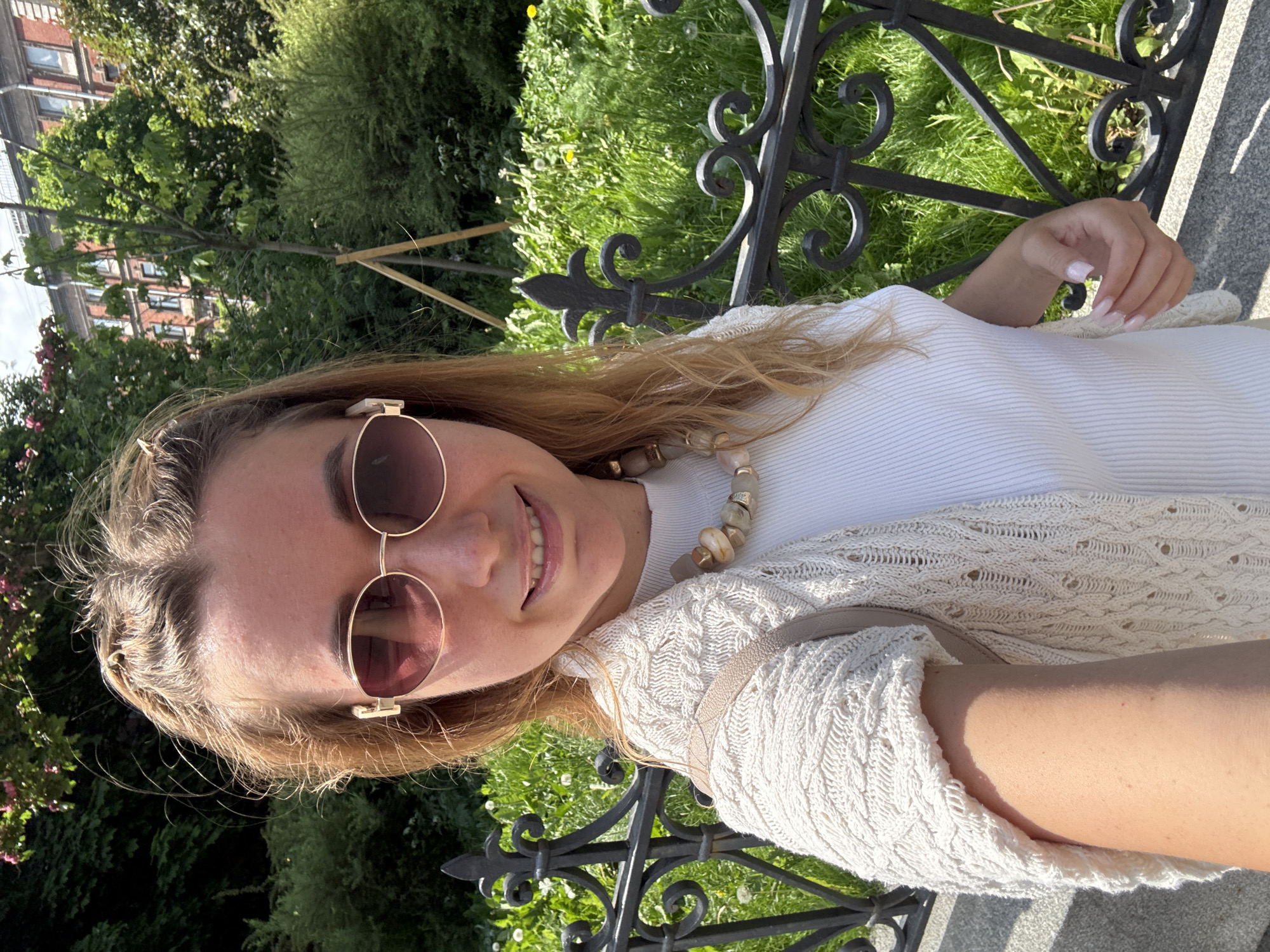 The image is oriented sideways, showing a close-up selfie of a smiling woman outdoors on a sunny day. The woman has long, light brown hair and is wearing large, round sunglasses with golden frames. She is dressed in a white ribbed top and a cream-colored, open-knit cardigan. A chunky necklace with light, irregularly shaped beads is visible around her neck. Her right hand, featuring manicured nails with light pink polish, rests near an ornate black metal fence.
The background consists of lush green foliage, including trees, bushes, and grass, suggesting a park or garden setting. In the upper left corner of the sideways image, part of a multi-story brick building with windows can be seen amidst the greenery, hinting at an urban or suburban environment. A grey stone or concrete surface is visible in the lower right, possibly a bench, path, or wall. The bright daylight and clear shadows indicate a sunny day. The woman is smiling broadly, conveying a cheerful and relaxed mood. No visible text is present in the image.
