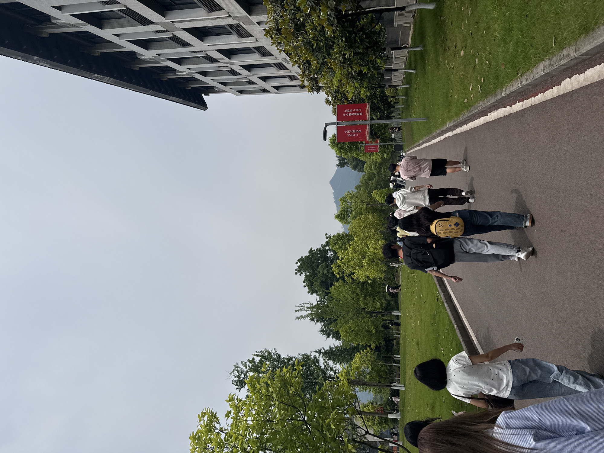 The image depicts a group of people walking along a paved path in Jiangjin, China. The path is bordered by a grassy area with trees on one side and a building on the other. In the background, a mountain is visible. Several red signs are seen near the path. The people are dressed in casual clothes and are walking in the same direction. One person appears to be holding a phone.