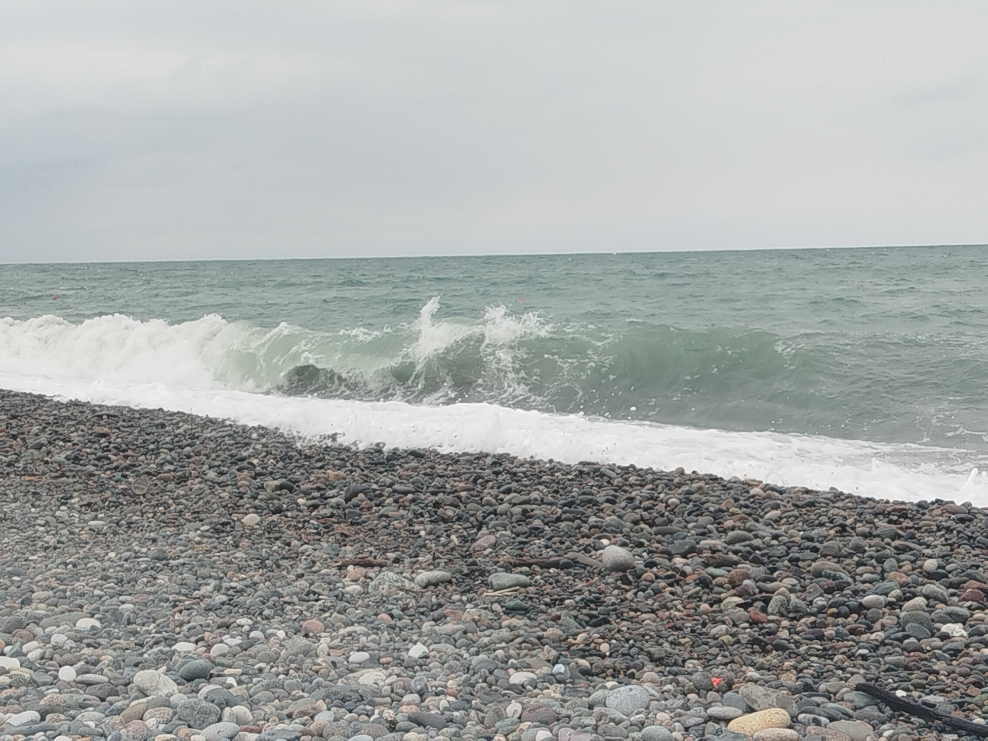 A pebble beach with a wave breaking in the distance is shown under a cloudy sky. The pebbles are a mix of gray, brown, and black, with some lighter colored ones interspersed. The water is a murky green, and the waves are white with foam. The sky is overcast and gray. The scene is calm and serene. It is likely the coast of Batumi, Georgia.