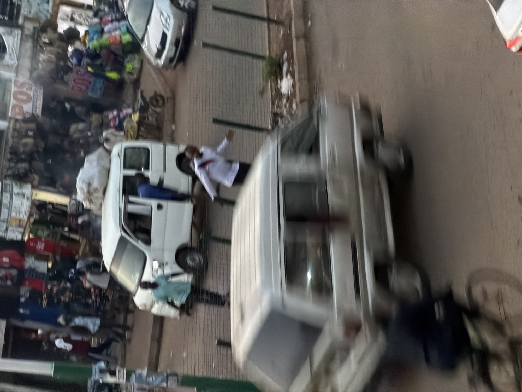 In Bukuru, Nigeria, on a dusty street, a street market is bustling with activity. A white minivan, with its doors open, is parked on the road, possibly for passengers. A man in a light blue shirt and dark pants stands beside the open door. Further down the road, a silver car is parked. In the foreground, a white minivan, its side facing the viewer, is in motion, blurred due to movement. A cyclist, also blurred, is riding on the left side of the road. The market stalls behind are filled with goods, and several people are seen browsing or working. The overall scene depicts a lively urban street in Nigeria.