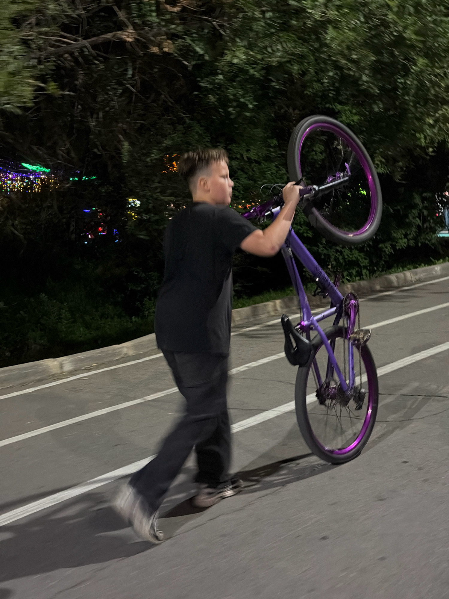 A young person with a dark t-shirt and pants is performing a wheelie on a purple BMX bicycle. The bicycle has purple rims. The action is taking place on a paved pathway with white lines, possibly a bike lane or pedestrian walkway. In the background, there are trees and some blurry, colorful lights that appear to be decorative, such as Christmas lights. The scene is dimly lit, suggesting it is evening or night. The location is specified as being in the city of Pavlodar, Kazakhstan. The overall impression is of an urban recreational activity at dusk or after dark.