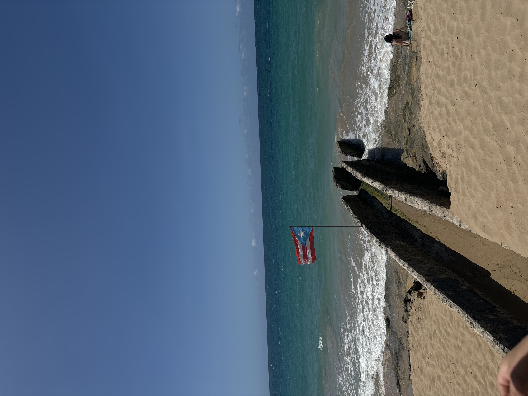 This is a medium shot of a beach in San Juan, Puerto Rico, on a sunny day. The Puerto Rican flag is flying on a pole to the left of a concrete structure that runs into the water. The ocean is turquoise with white waves crashing on the shore. A person is sitting on the rocks by the water to the right of the concrete structure, facing away from the camera. There are a few items of clothing or towels on the sand next to the person. The sand is golden and there are some rocks scattered on the beach. The sky is clear and blue.