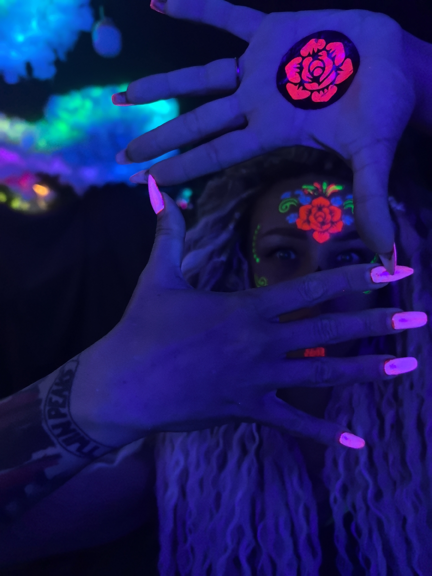 A person with blonde hair is depicted under blacklight, creating a vivid, colorful scene. The person has a floral design painted on their forehead, specifically a red rose surrounded by green and blue accents. The person's hands are positioned in front of their face, framing their eyes. On the back of one hand, a similar red rose design is present. The person has long, pink-glowing nails. A tattoo is visible on their arm. The background is dark with some brightly illuminated shapes in the upper left. The specific location of Noviy, Russia is not discernible.