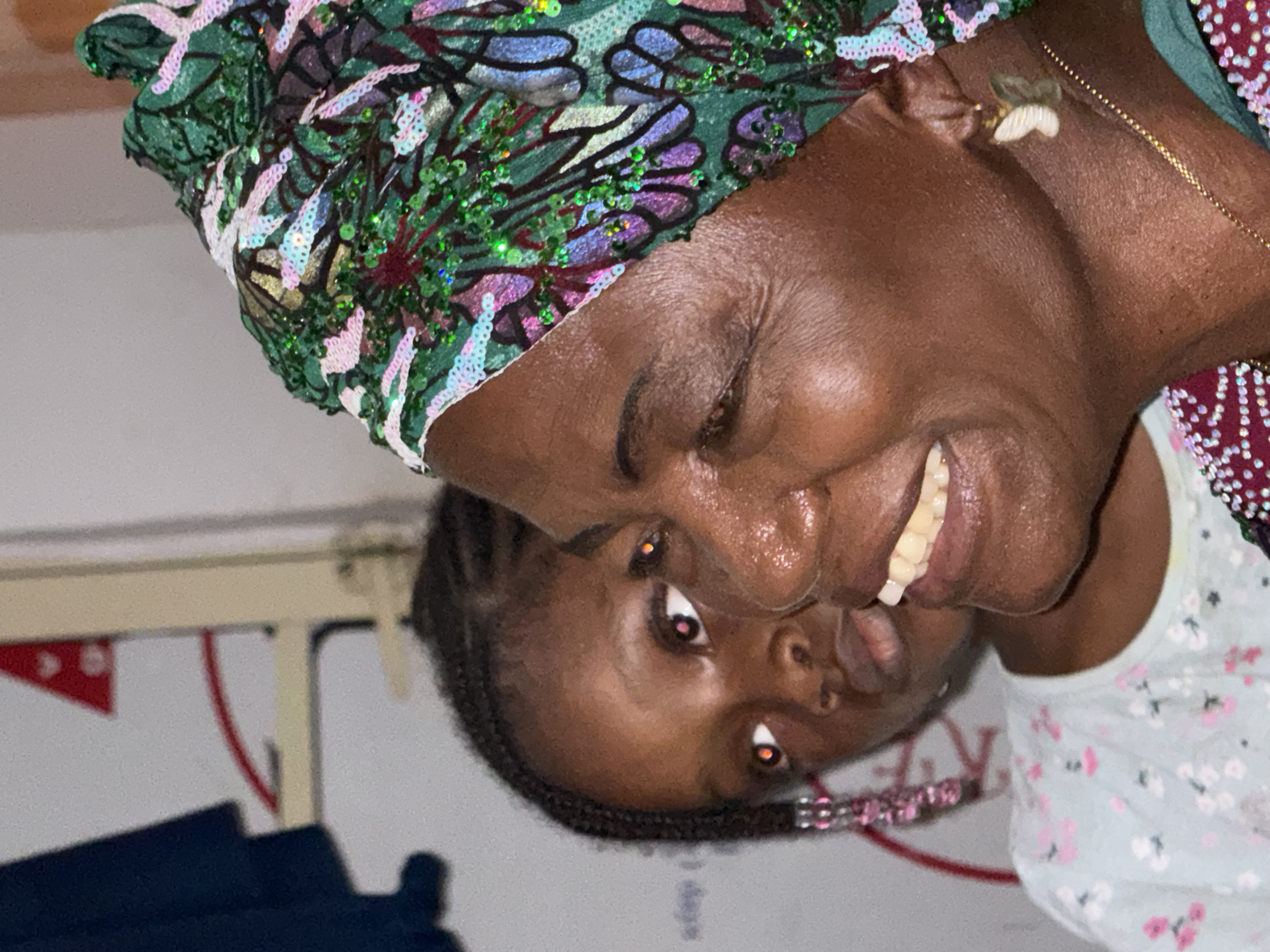 A medium shot of a woman wearing a sequined green, white, and purple head covering, wearing gold jewelry, smiling, and standing next to a child with cornrow braids, likely indoors, in Ado, Nigeria.