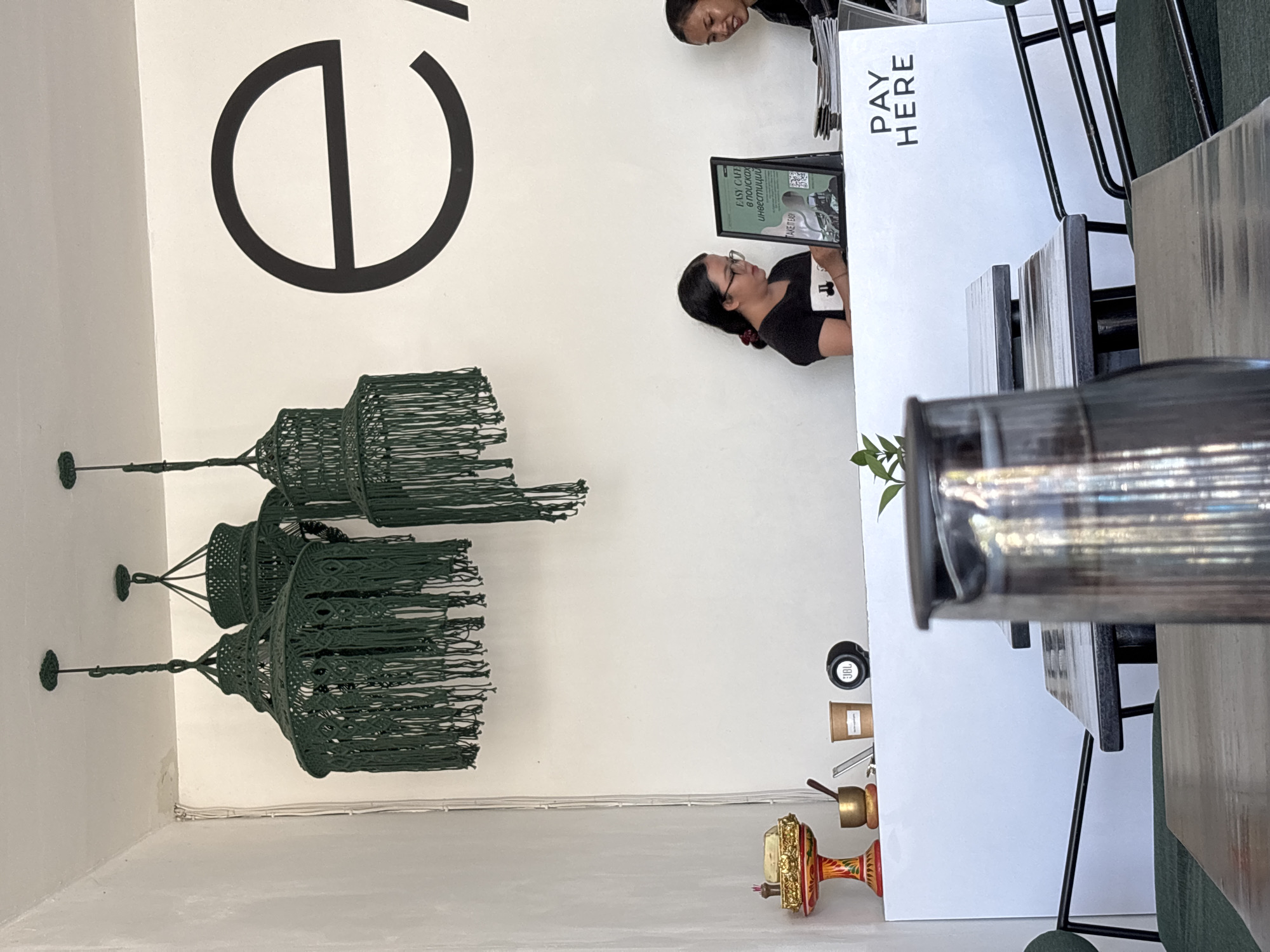 An interior shot depicts a casual commercial space, likely a cafe or retail shop, featuring a checkout counter and decorative elements. A woman with dark hair and glasses is positioned behind a white counter, holding a cup and looking intently at a digital display. The head of another person is partially visible behind and above her, also facing the counter area.
The background consists of a plain white wall, adorned with a large, black, stylized circular logo containing a vertical line. To the left, two prominent dark green macrame hanging light fixtures or decorations are suspended from the ceiling.
On the white counter, a clear sign reads "PAY HERE". A black digital display or tablet stands upright on the counter in front of the woman, showing text that includes "EASY CAFE", Greek words such as "εποχικα" and "επισκοπη", and "TAKE IT TO", alongside a QR code. On a lower shelf or adjacent surface, a black JBL speaker, a small brown box, and a decorative, colorful brass-like bell or mortar are arranged. A small green plant adds a touch of nature near the counter's edge. The scene suggests a transaction or interaction taking place at a point of sale, set in Kuta Selatan, Indonesia.