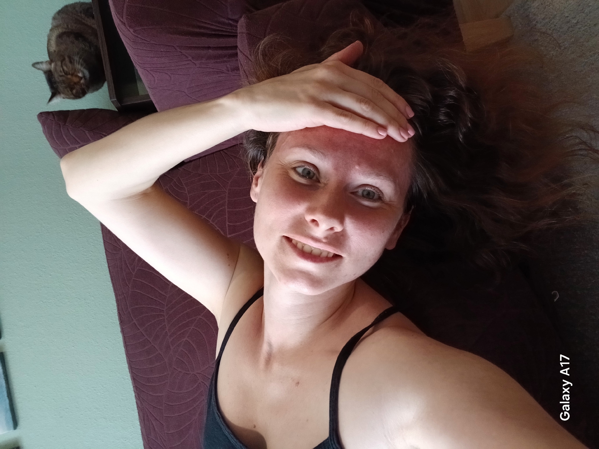 A woman is lying on a sofa with her head tilted back and her arm resting on her forehead. She is wearing a black tank top. A cat is sitting on a shelf in the background. The room appears to be a living room, with a green wall and a brown sofa. The image was taken with a Galaxy A17 phone. There is no information in the image that indicates it was taken in Yekaterinburg, Russia.