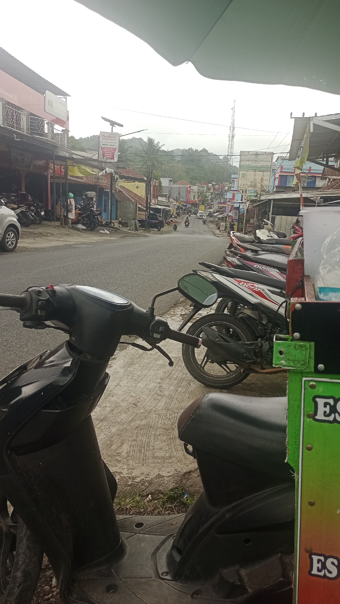 A street scene in Karyamandala, Indonesia, features a row of parked motorcycles along the side of a road. The road itself is paved and shows some wear. Buildings line the street, appearing to be shops or businesses, with signage visible on some. In the background, there are more buildings and a hilly landscape under an overcast sky. A telecommunications tower is visible in the distance. The foreground is dominated by the front of a black scooter. The overall impression is that of a typical street in a town or village in Indonesia, with abundant motorcycle traffic and commerce.