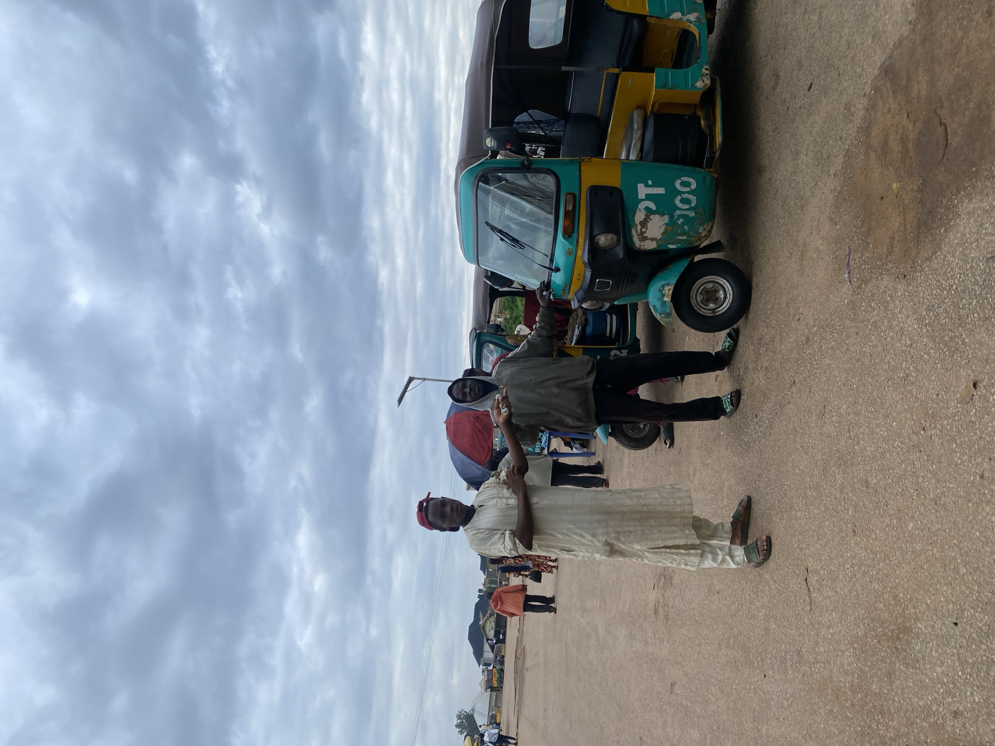 In Rafin Par, Nigeria, two men stand in front of two auto-rickshaws under a cloudy sky. The man on the left, wearing a light-colored, striped robe and a red baseball cap, holds a small object in his right hand, extended towards the man on the right. The man on the right, dressed in a dark hooded jacket and dark pants, also holds a small object in his hand. Both men appear to be interacting, possibly exchanging the items. The auto-rickshaws, painted in teal and yellow, are parked on a dusty road. In the background, other people and structures suggest a bustling area. The overall scene captures a moment of exchange in a busy Nigerian town.
